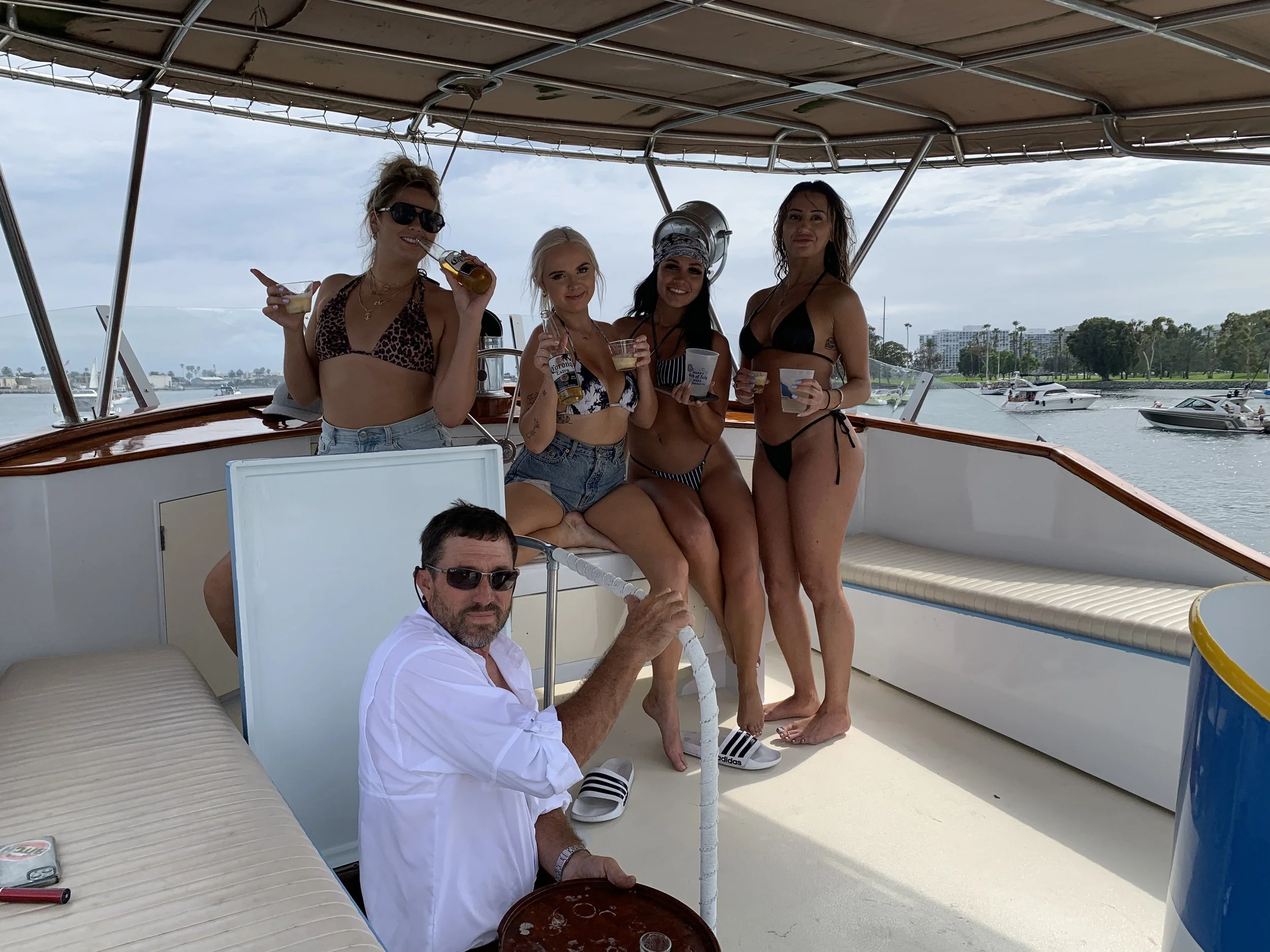 Group of five friends on a boat enjoying drinks, with some wearing swimsuits, surrounded by water and boats in the background.