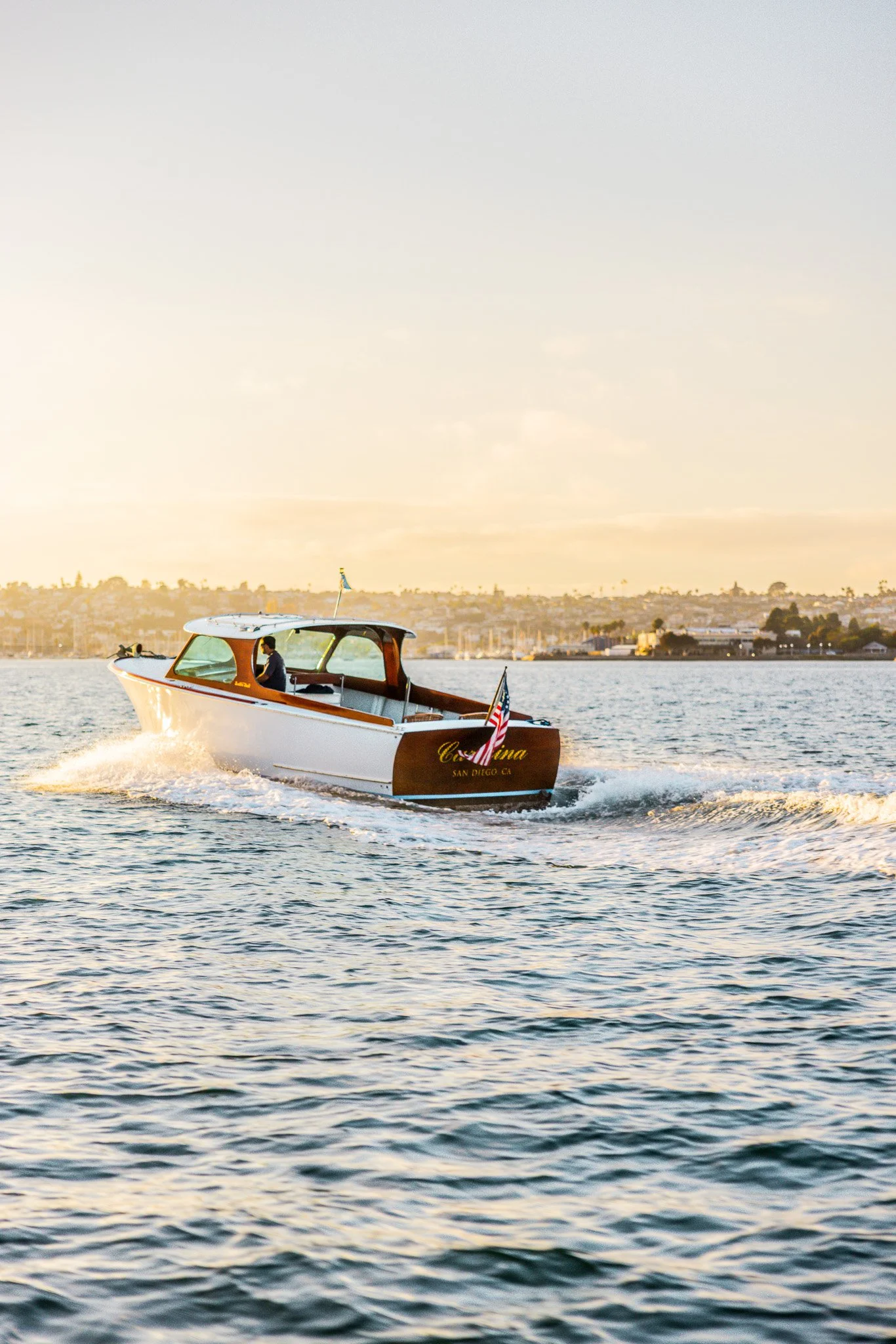 A white motorboat with wooden accents cruising on the water during sunset, with a cityscape in the background.
