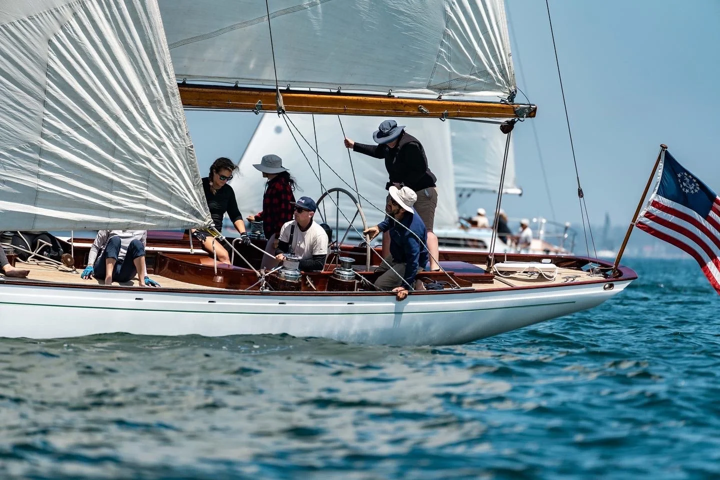 A group of people sailing on a boat with a large white sail, some wearing hats and sunglasses, navigating through the water, with an American flag on the boat's stern.