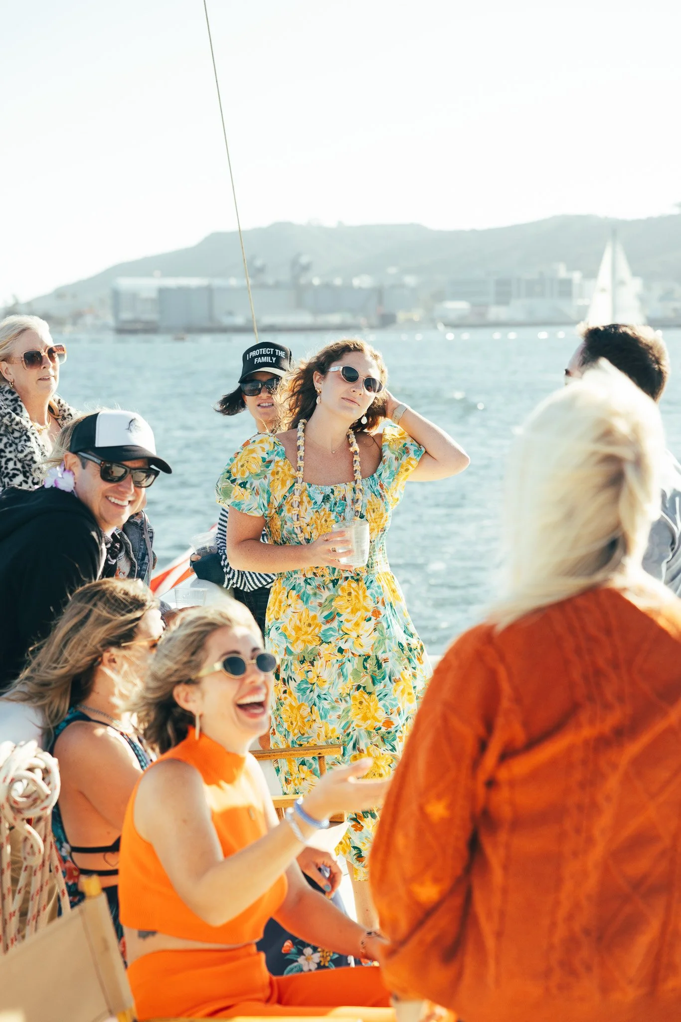 Group of women on a boat enjoying a sunny day, some wearing sunglasses and colorful summer dresses, with water and cityscape in the background.