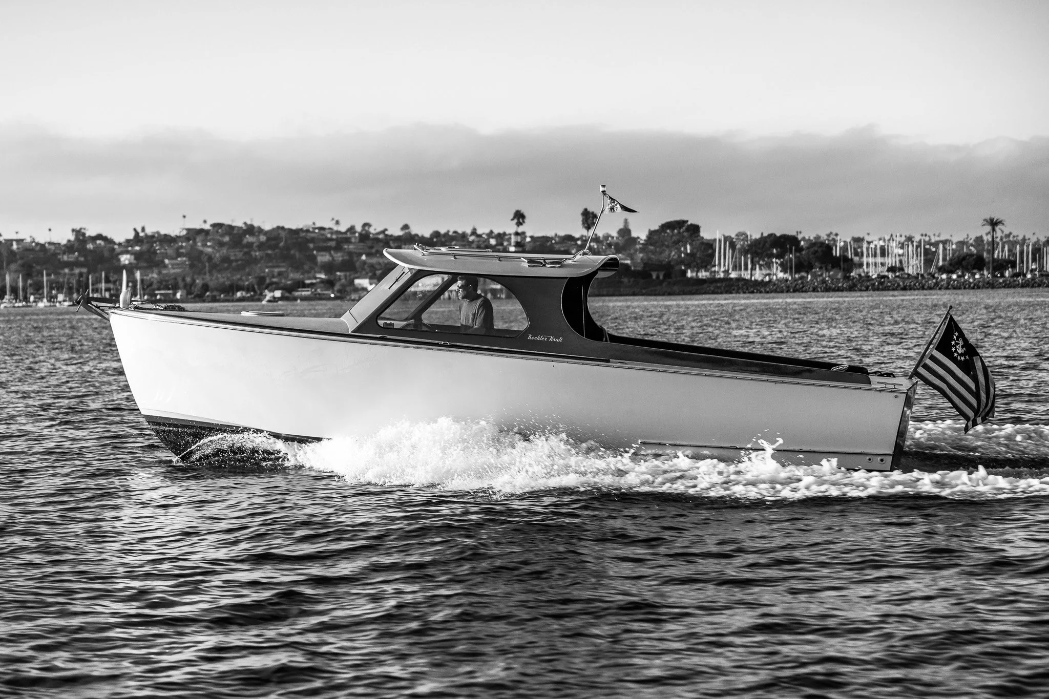 A person on a motorboat moving through water, with a city shoreline and sailboats in the background, shown in black and white.