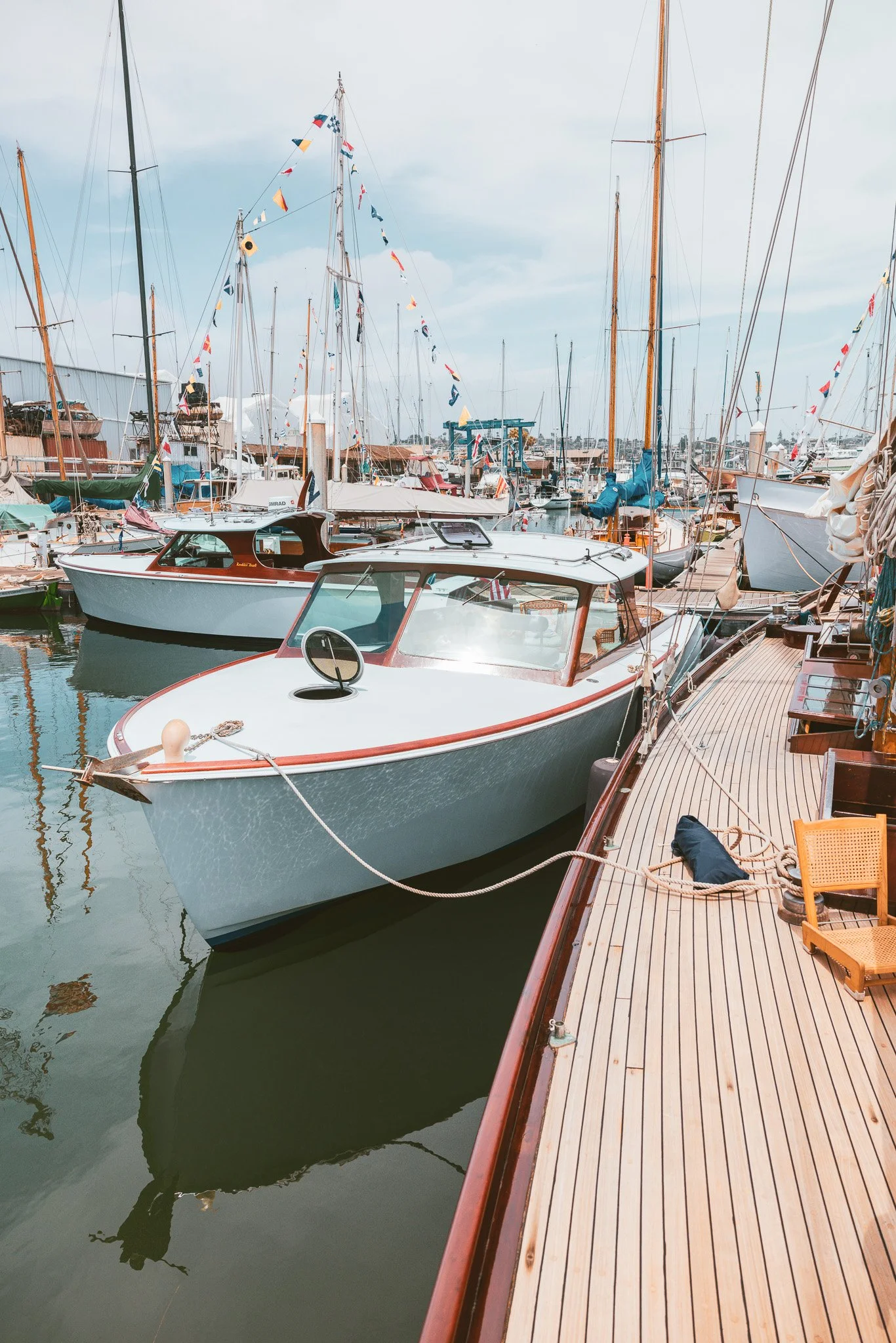 A marina filled with anchored sailboats and motorboats, with colorful flags hanging from the masts, alongside a wooden dock.