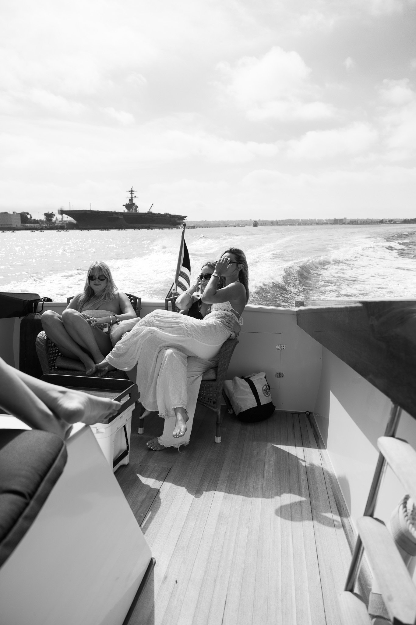 Three women sitting on a boat with a wake behind; a warship visible in the water; women in casual attire, one with sunglasses, relaxed during daytime.