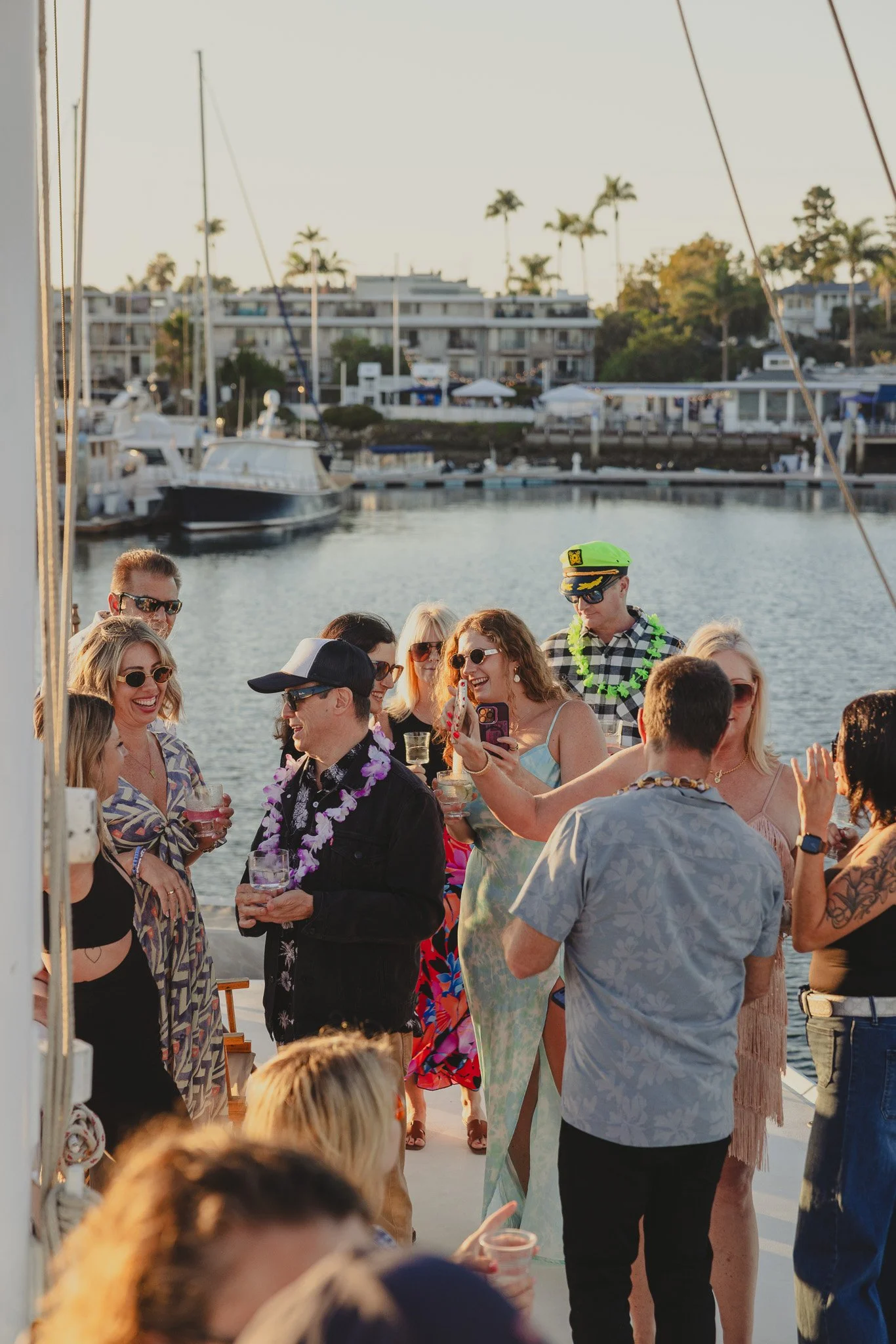 People celebrating on a boat docked by a marina during sunset, wearing leis, with some holding drinks and taking pictures, in a festive outdoor setting.
