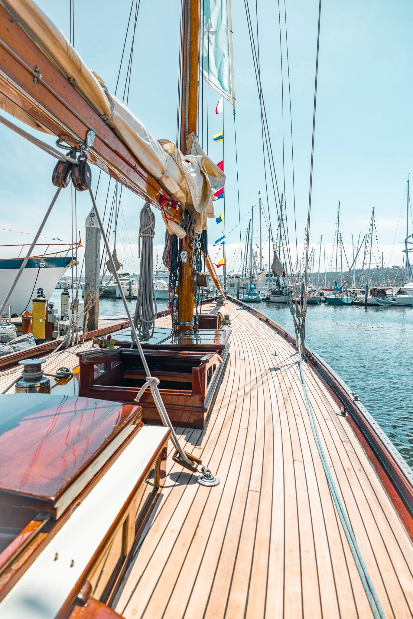 A view from the deck of a wooden sailboat docked at a marina, with several other boats and masts in the background under a clear blue sky.