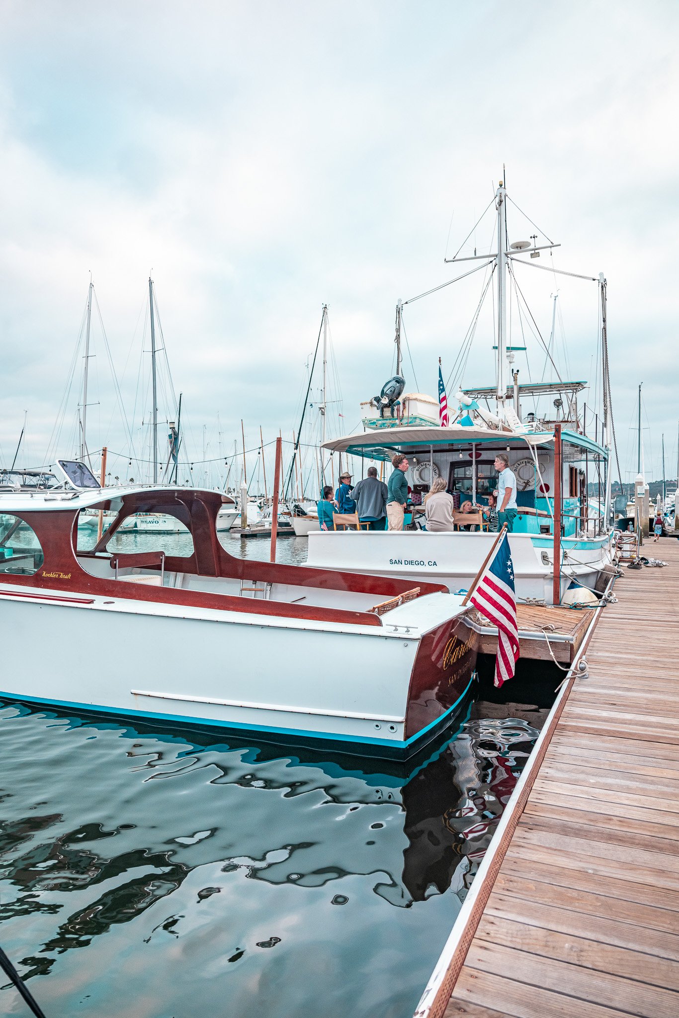 Boats docked at a marina in San Diego, California, with people gathered on the main boat, and a wooden pier on the right side.