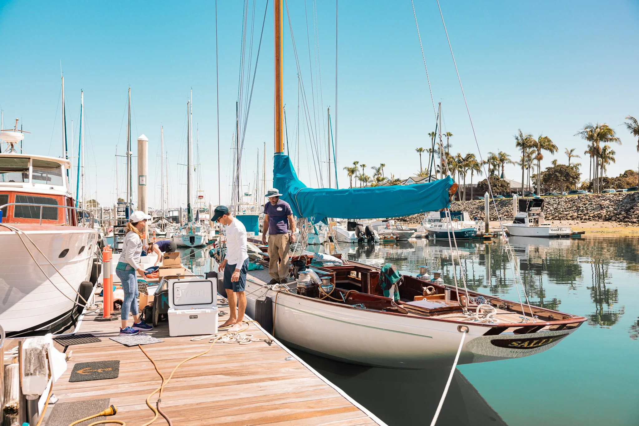 People preparing a sailboat at a marina dock on a sunny day with clear blue skies, surrounded by other boats and palm trees in the background.