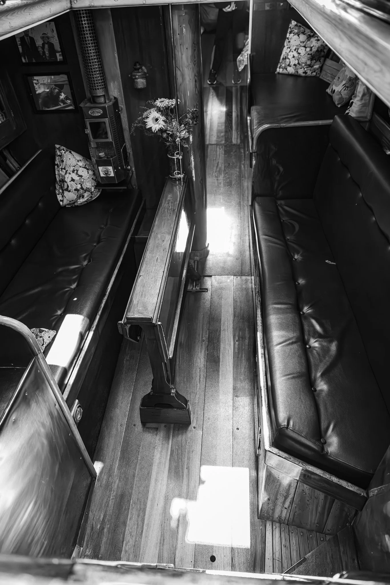 Interior of a vintage bus with black leather bench seats, wooden flooring, photographs and flowers on the walls, and a small table between the seats.