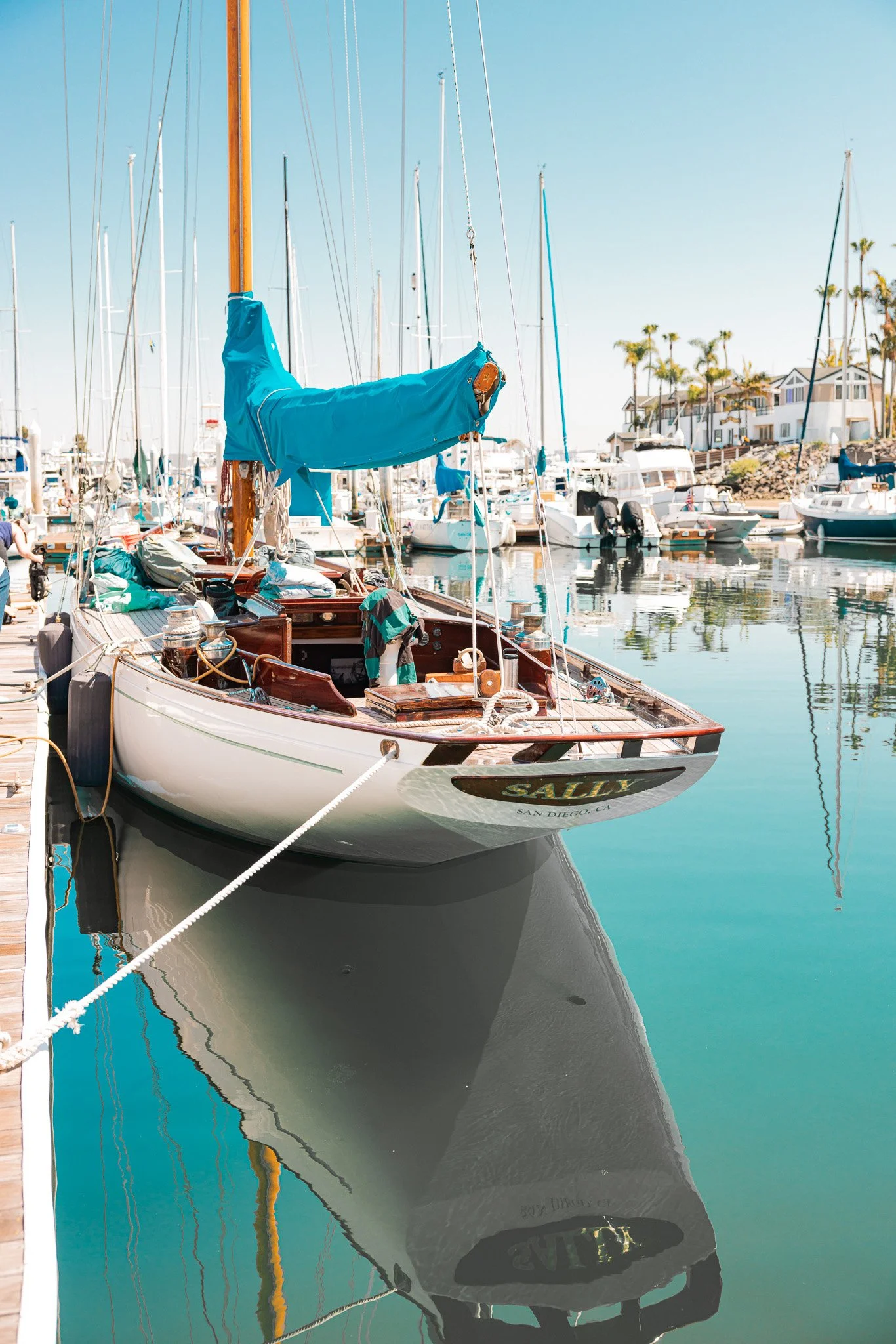 A sailboat docked at a marina with calm blue water, other boats, and houses with palm trees in the background on a sunny day.