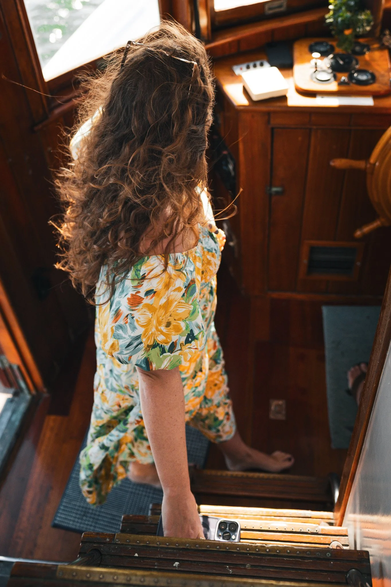 A woman with long, curly brown hair wearing a floral dress walking upstairs inside a wooden boat or cabin, holding a smartphone in her hand.