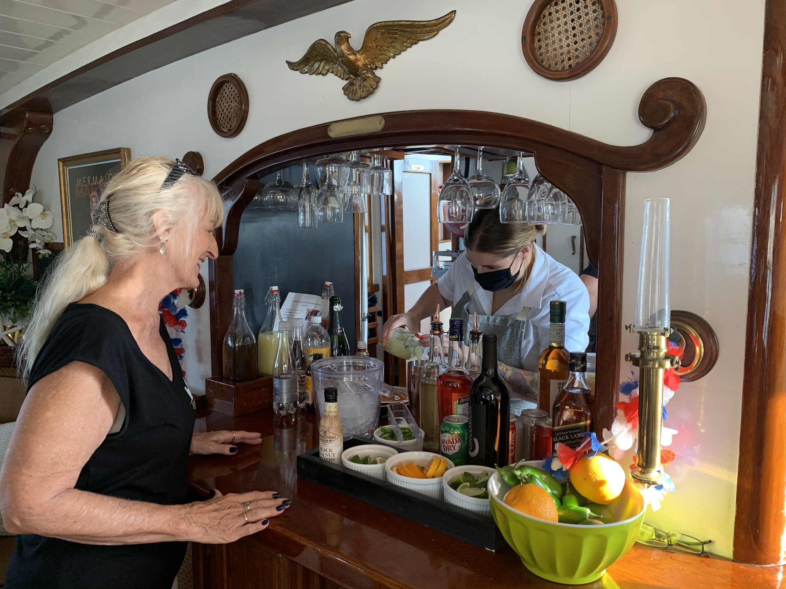 Older woman with long white hair smiling and standing at a bar, talking to a bartender. The bartender, wearing a black face mask and a white shirt, is preparing a drink behind the bar. The bar is decorated with bottles of alcohol, bowls of fruit, and