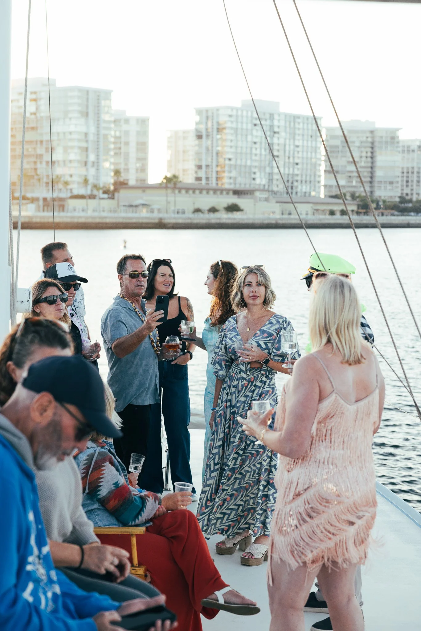 Group of people enjoying a boat party by the water, with city buildings in the background. Some are holding drinks and taking photos, dressed in casual and summer attire.