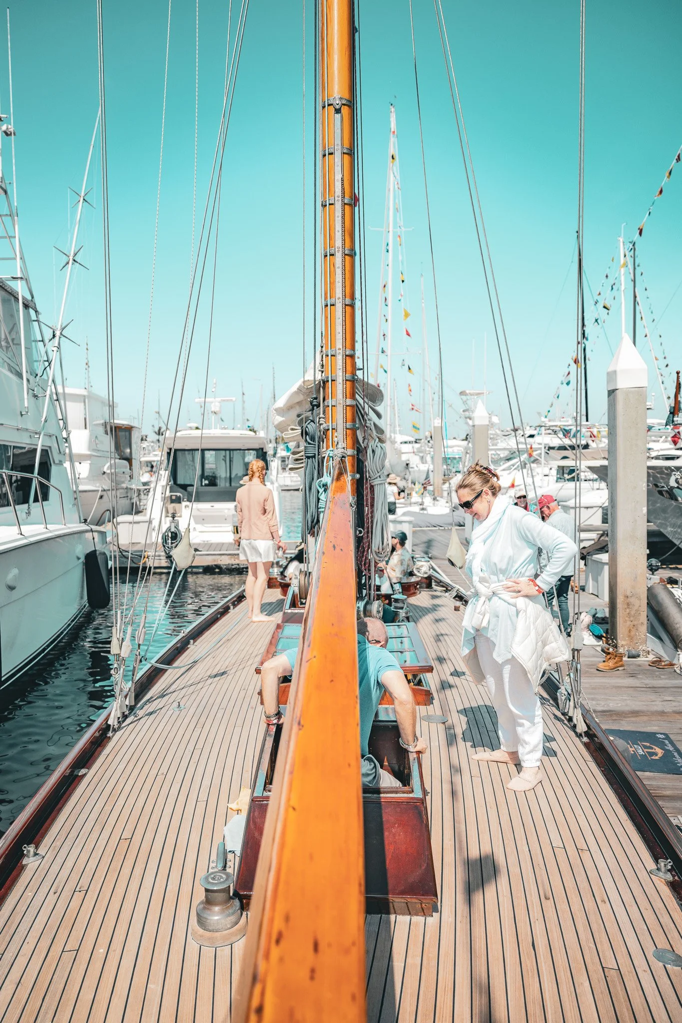 People on a wooden boat docked at a marina with lots of other boats, some people are sitting and others are walking, the sky is clear and blue.
