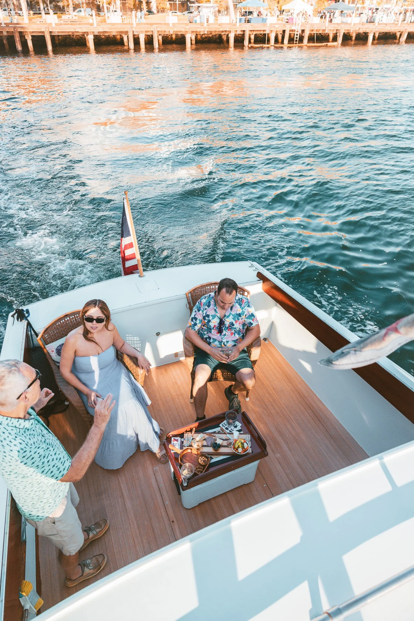 Three people on a boat enjoying food and drinks, with water and a dock in the background.