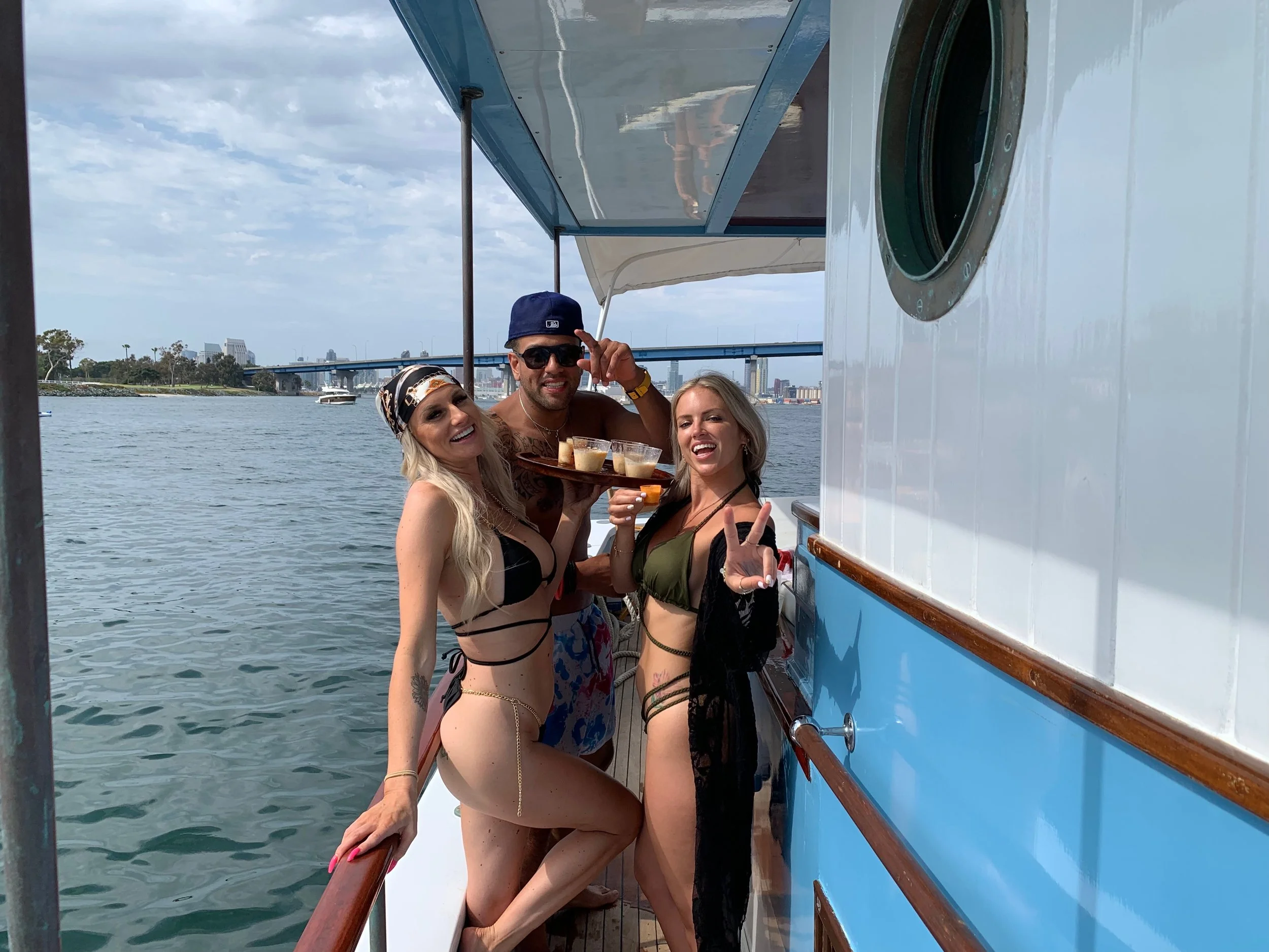 Three friends in swimsuits on a boat, holding drinks and smiling at the camera, with a city skyline in the background.