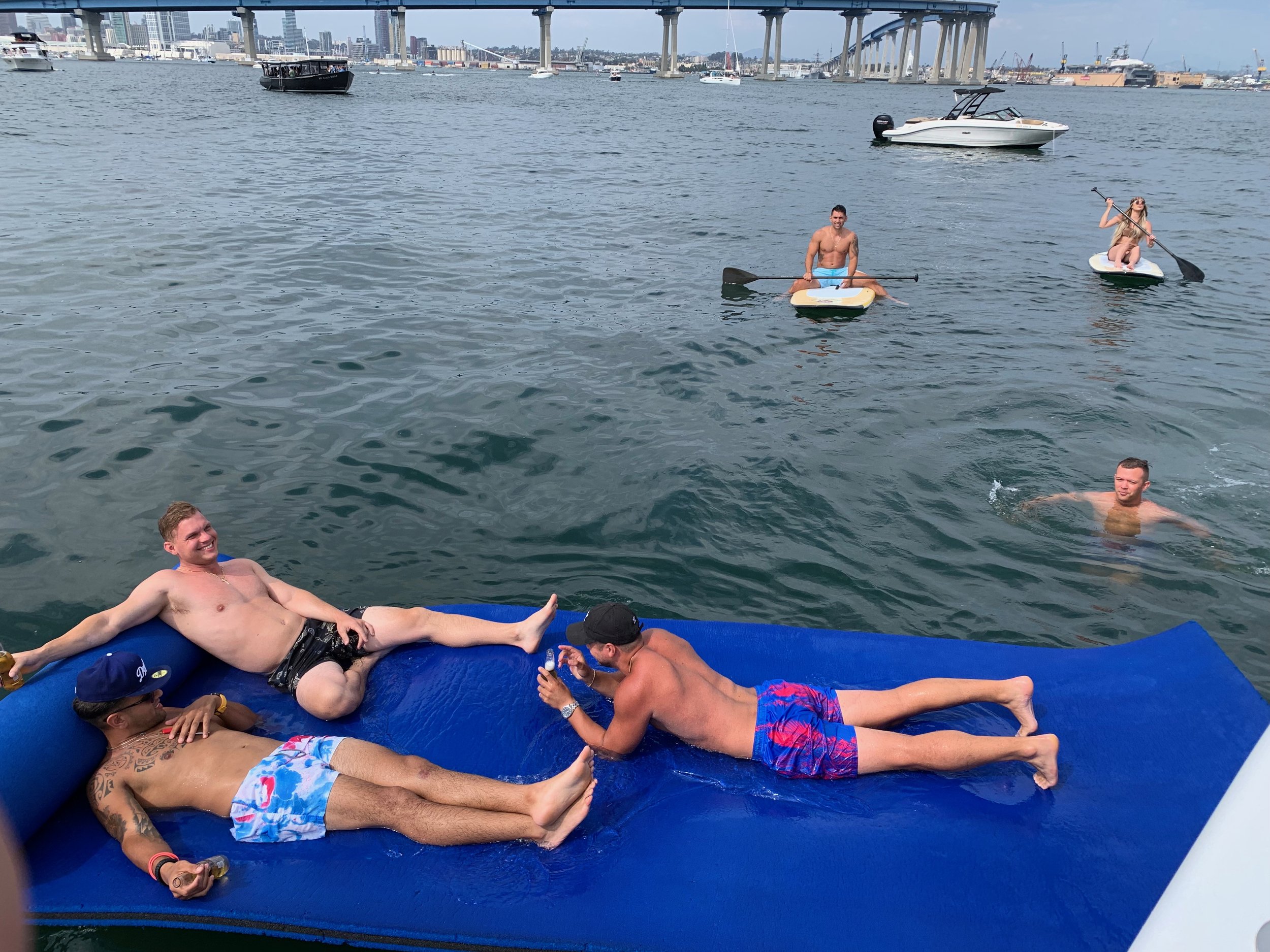 Group of friends relaxing and swimming in a harbor with boats and a bridge in the background, some on a floating mat and others on paddleboards.