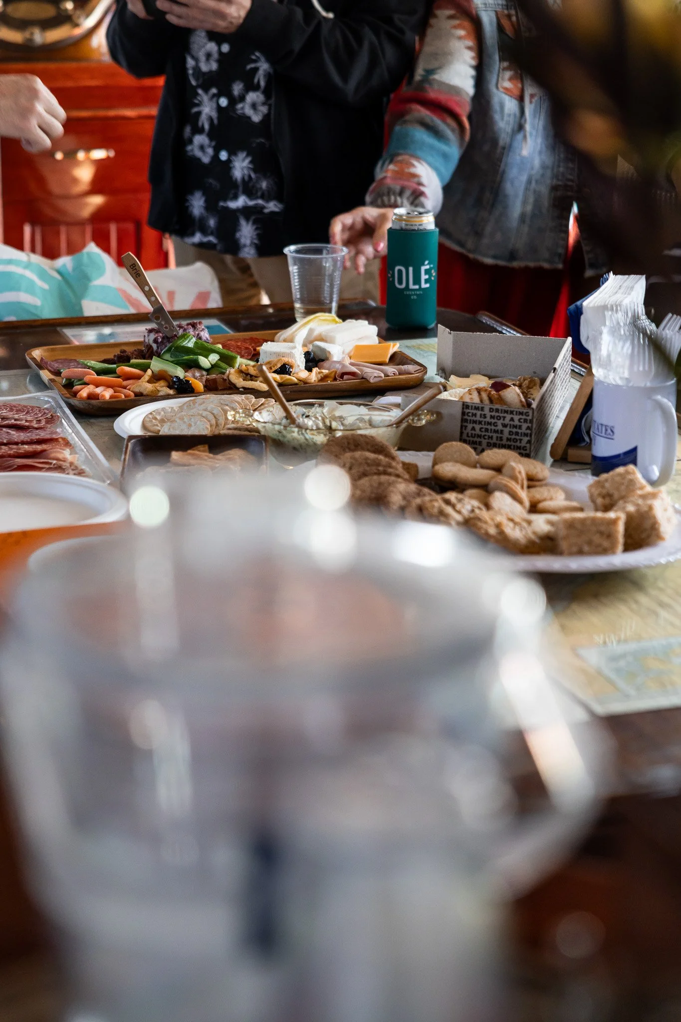 A table filled with various food and drink items, including cheese, crackers, vegetables, and meat, with people standing around it.