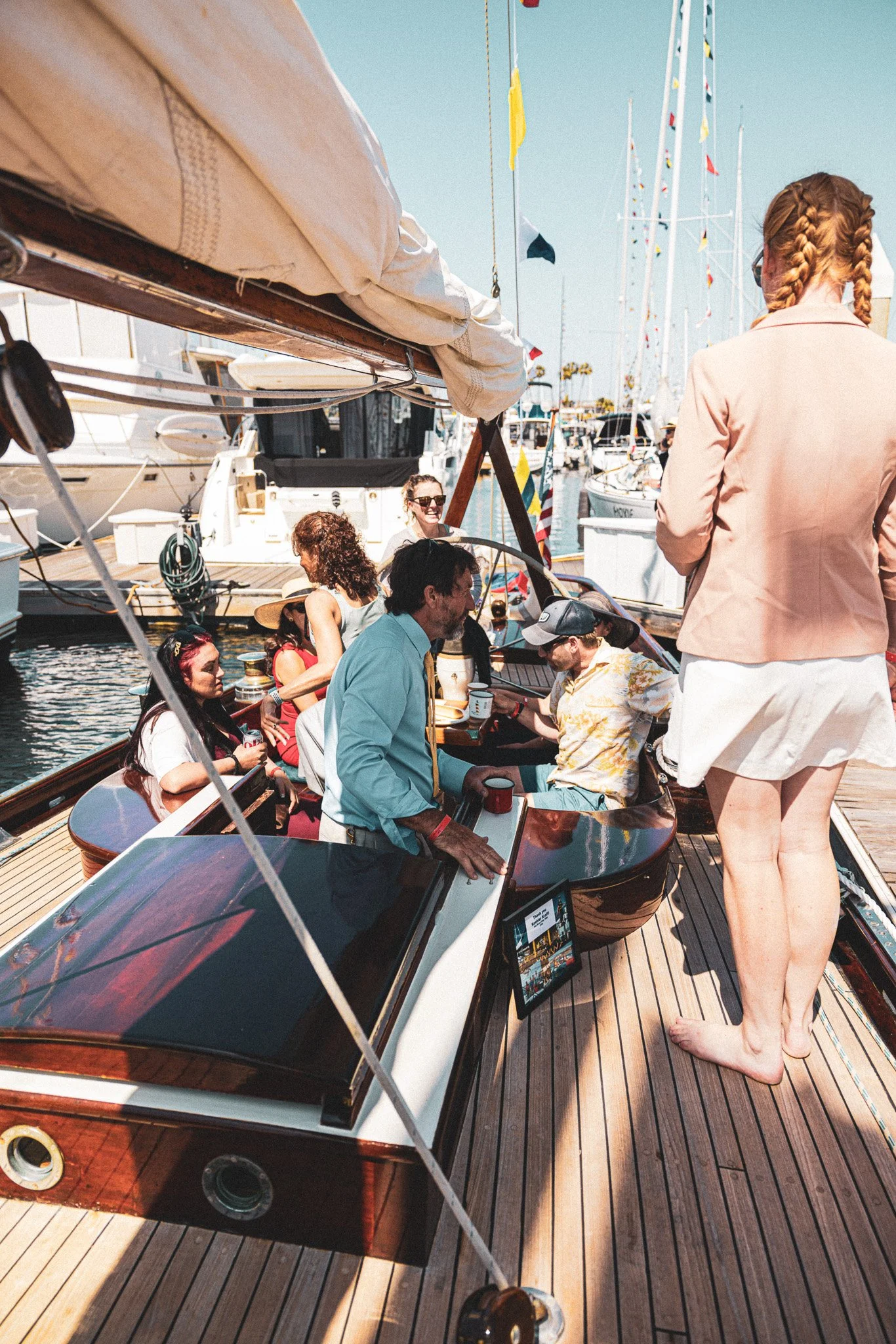 Group of people enjoying their time on a yacht docked at a marina with multiple sailboats and American flags in the background.