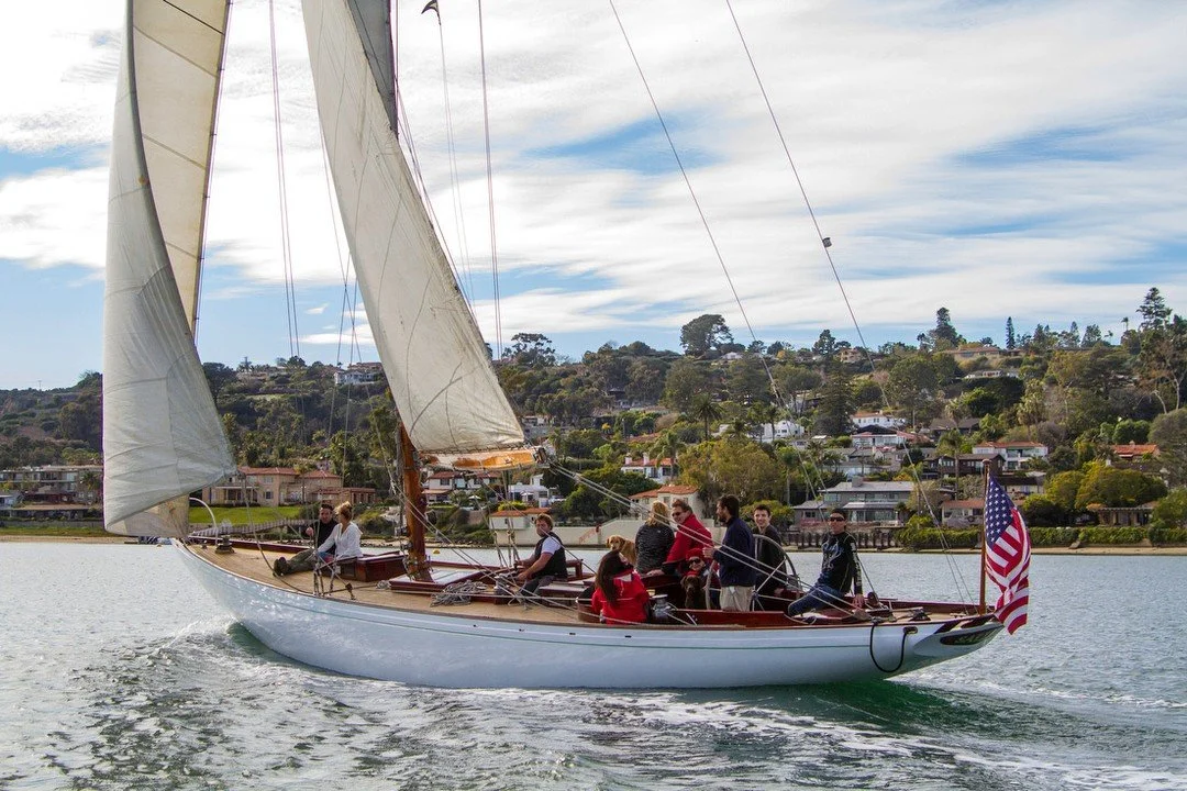 A sailboat with white sails sailing on a lake with a group of people onboard, with houses and trees on a hillside in the background.