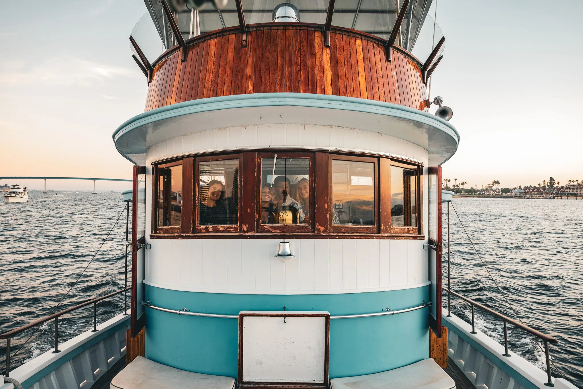 People smiling inside a boat's observation deck, seen through windows with a waterfront and bridge in the background during sunset.