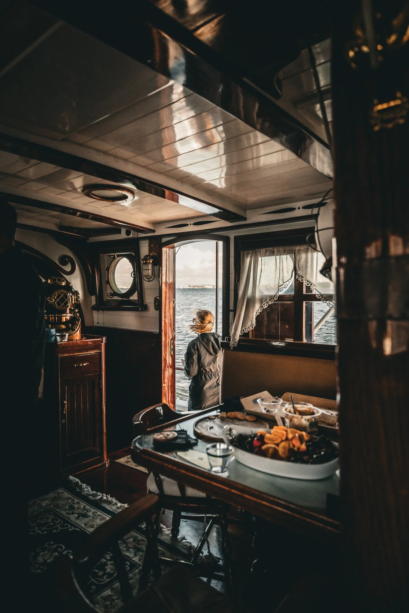 A person standing at the doorway of a boat looking out at a body of water, with breakfast or snacks on a table inside the boat.