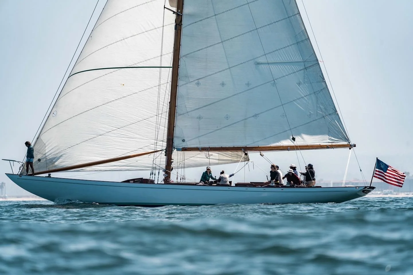 Sailboat with multiple sails and several people onboard, sailing in open water, with American flag at the stern.