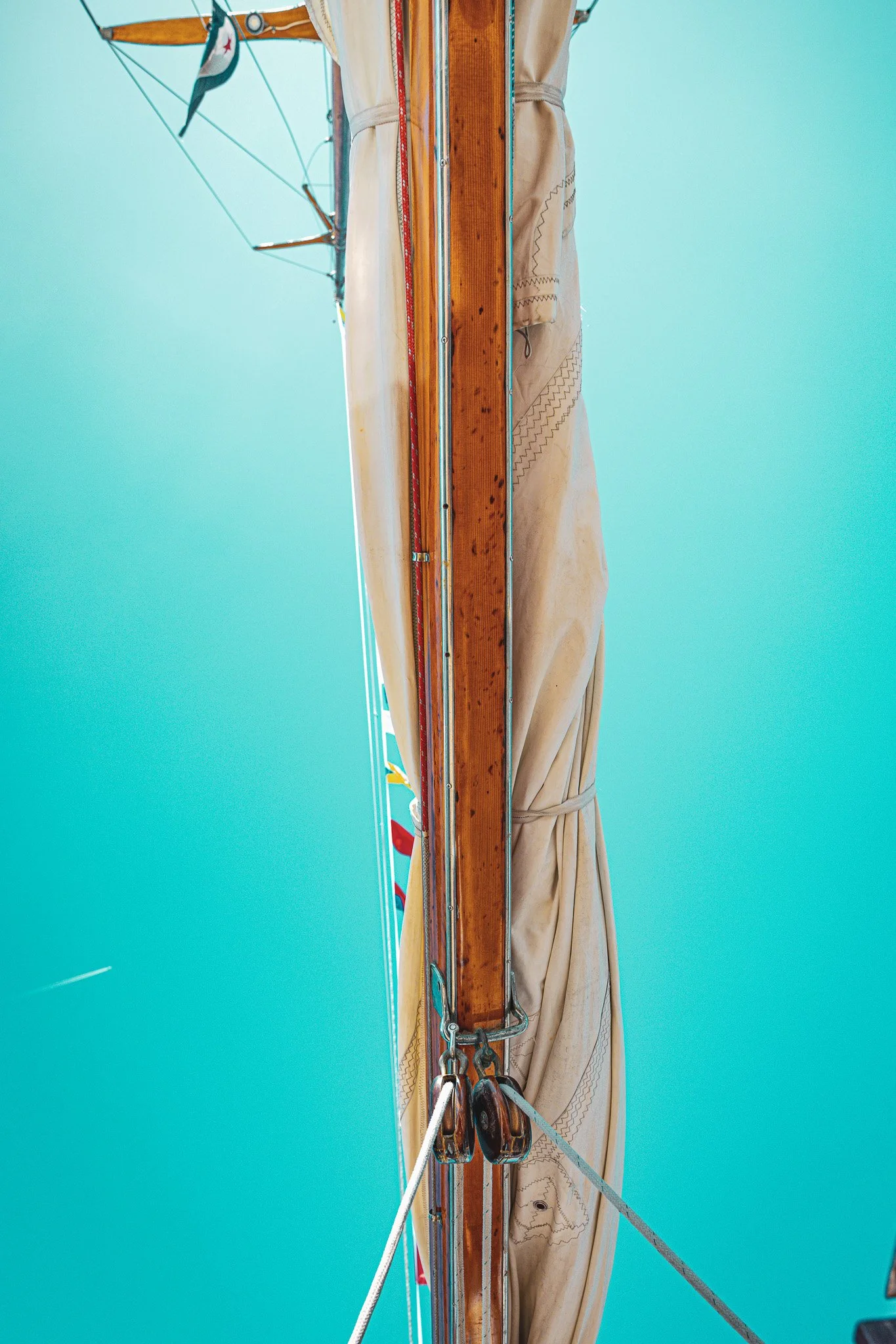 Close-up view of the mast of a sailboat with beige sails furled, reaching up toward a clear blue sky.