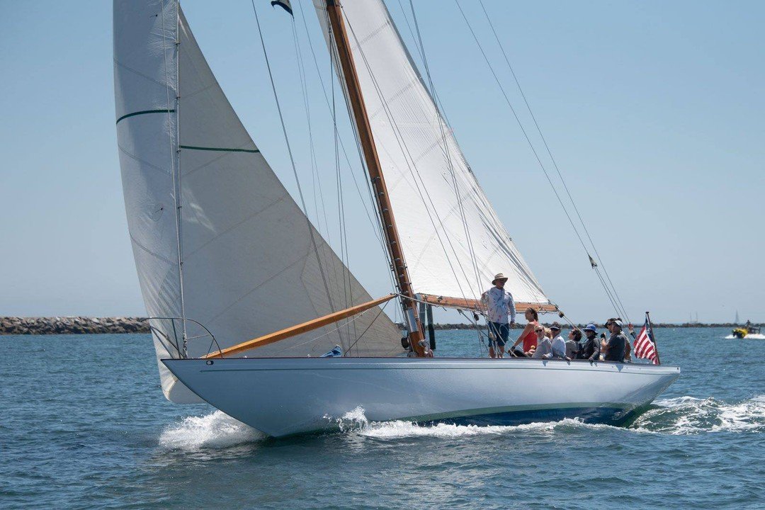 A sailboat with multiple sails on the water, carrying several people, with a breakwater in the background and a clear blue sky.