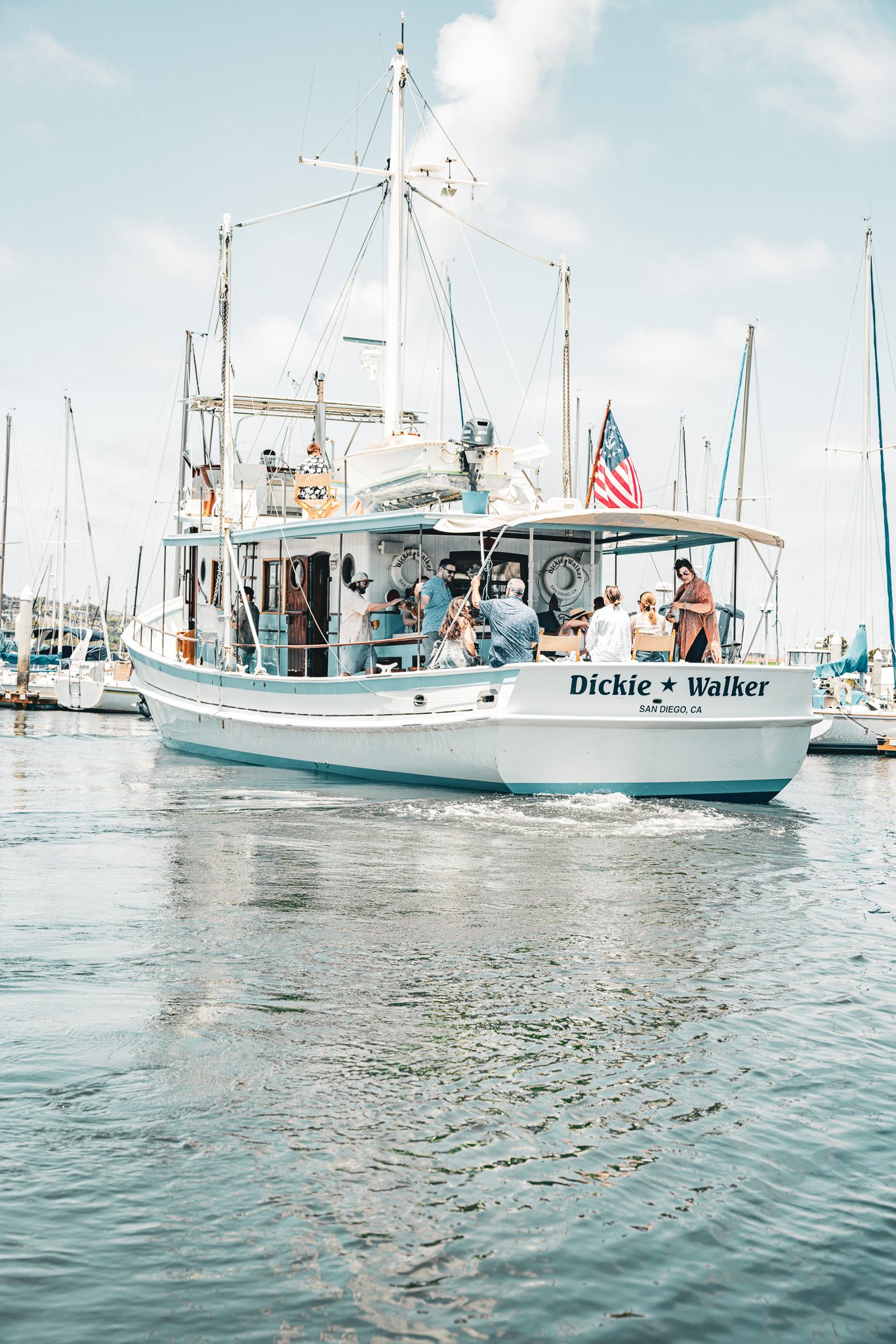 People enjoying a boat ride on a white yacht named Dickie Walker on a marina with multiple boats and sailboats in the background