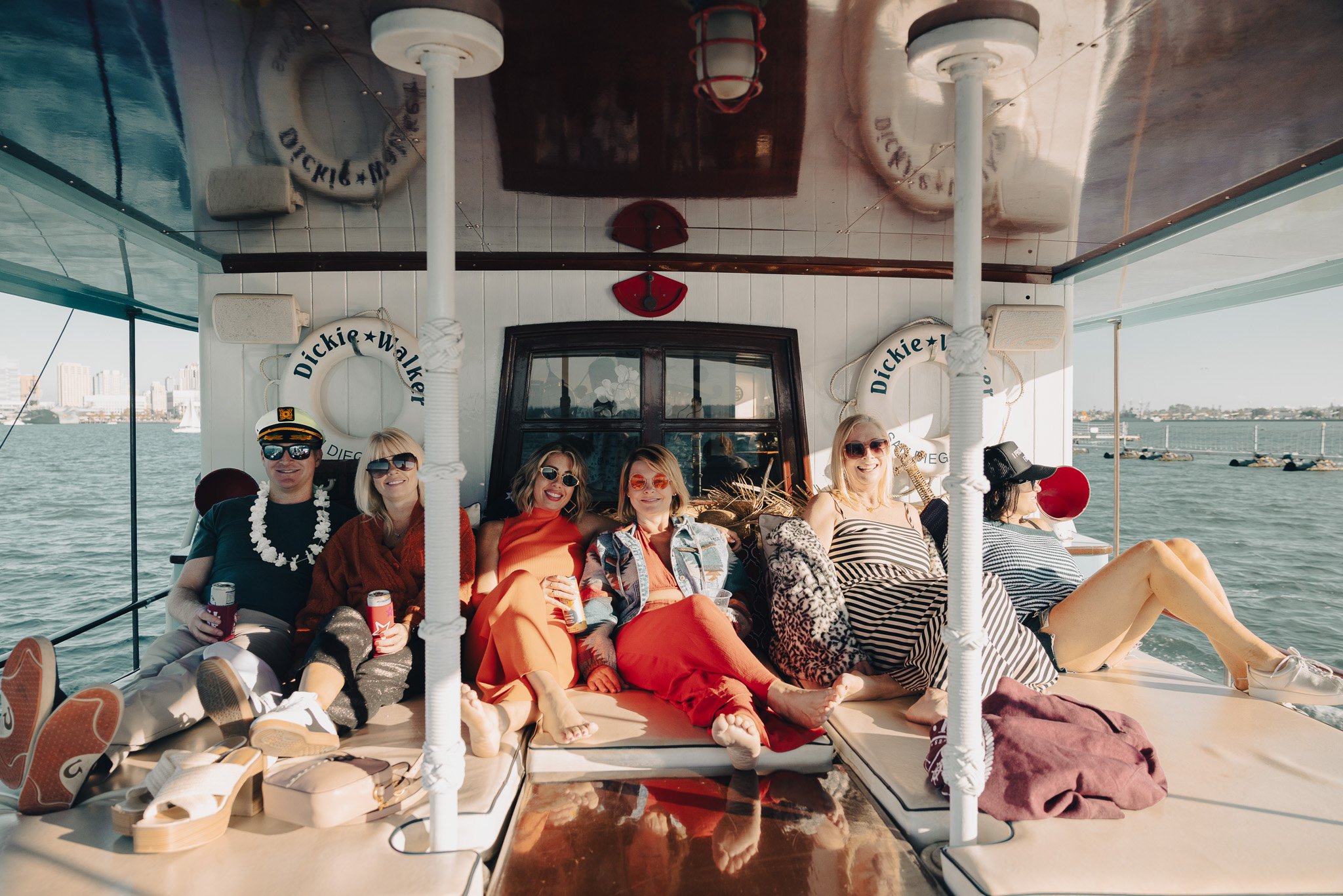 Group of six friends relaxing on a boat, sitting on cushions, dressed in casual summer clothing, with water and city skyline visible in the background.