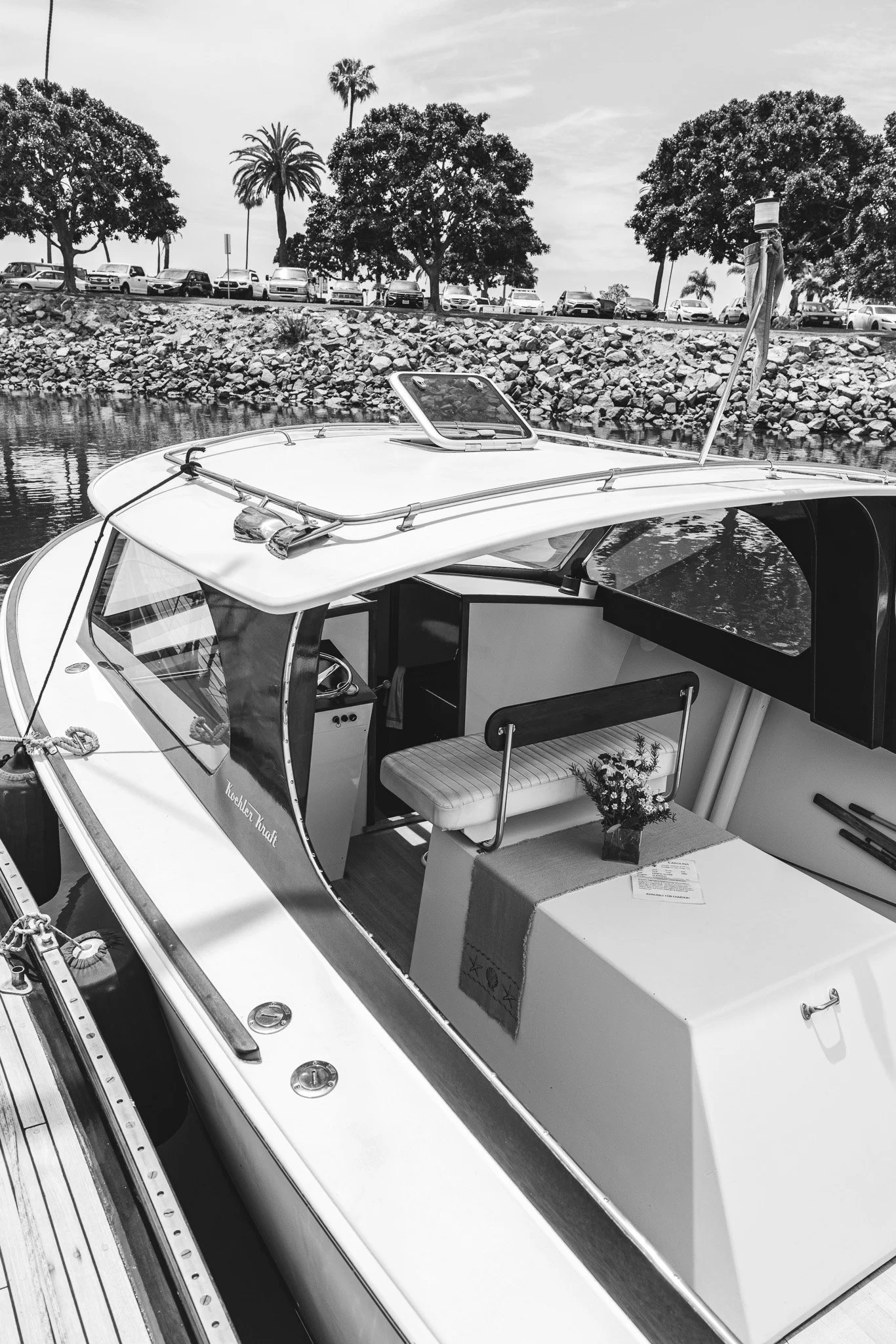 Black and white photo of a boat docked at a marina with trees and parked cars in the background.
