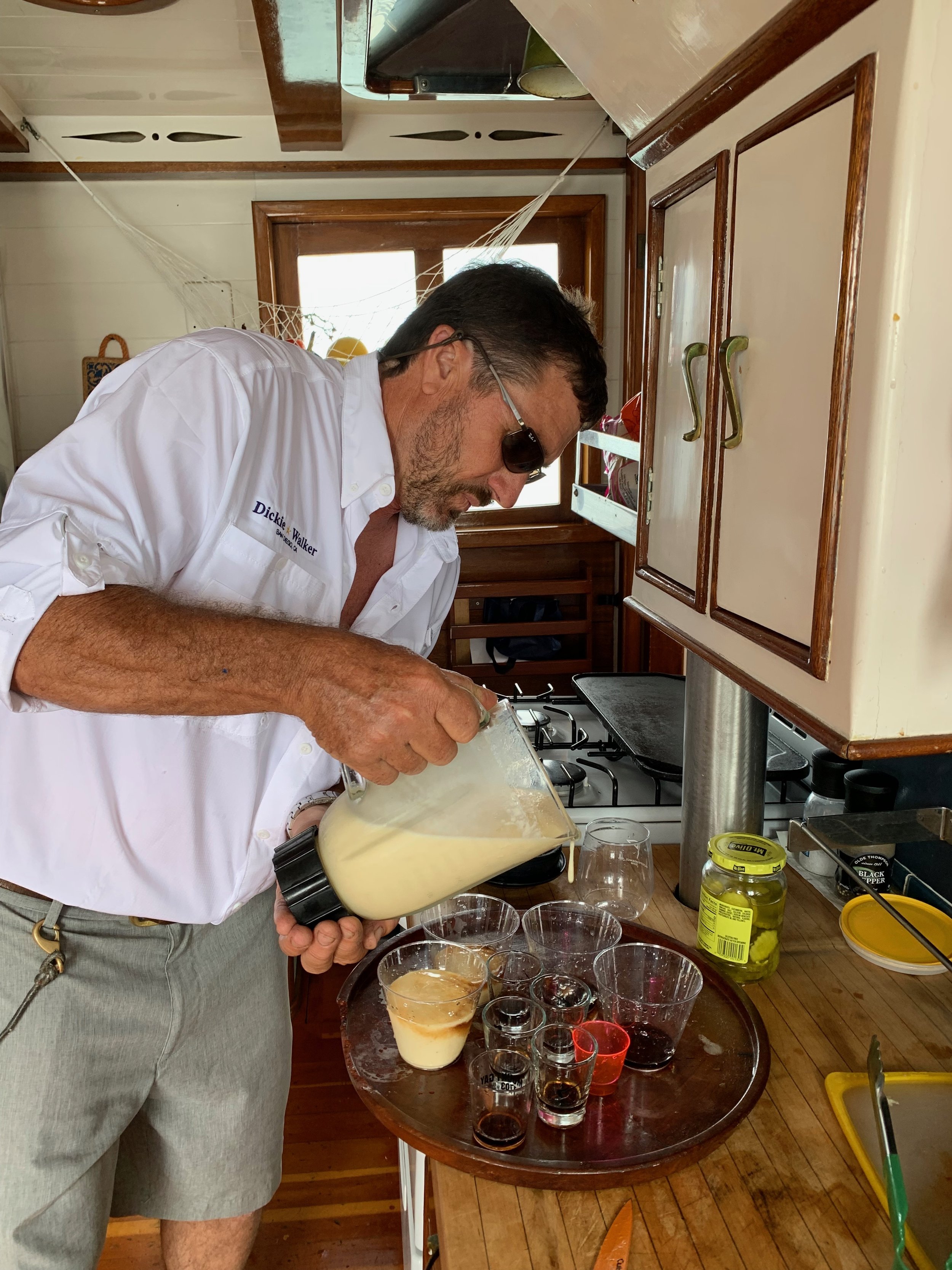 A man in sunglasses and a white shirt pouring a creamy drink into small glasses on a wooden table, with various beverages and jars nearby, in a rustic kitchen.
