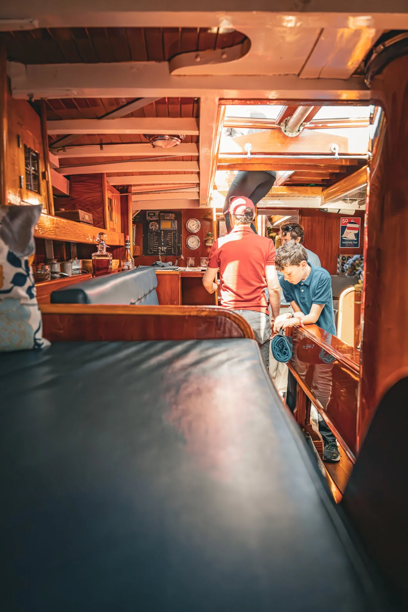 Inside a wooden boat with people gathered, some standing and some sitting, in a cozy, sunlit space with nautical decor.