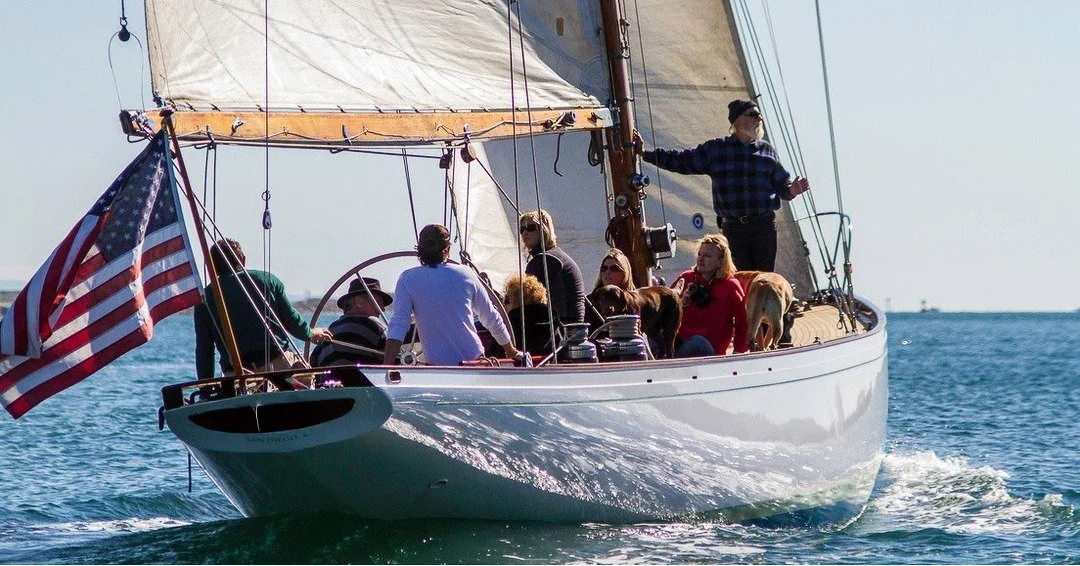 A group of people and a dog sailing on a white sailboat with an American flag, on the water during the day.