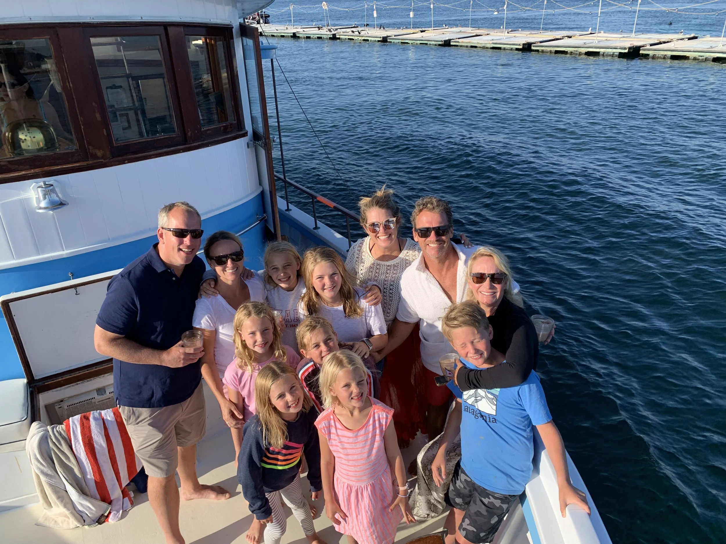 Family and friends on a boat celebrating, holding drinks, smiling, and enjoying a sunny day on the water with a dock in the background.