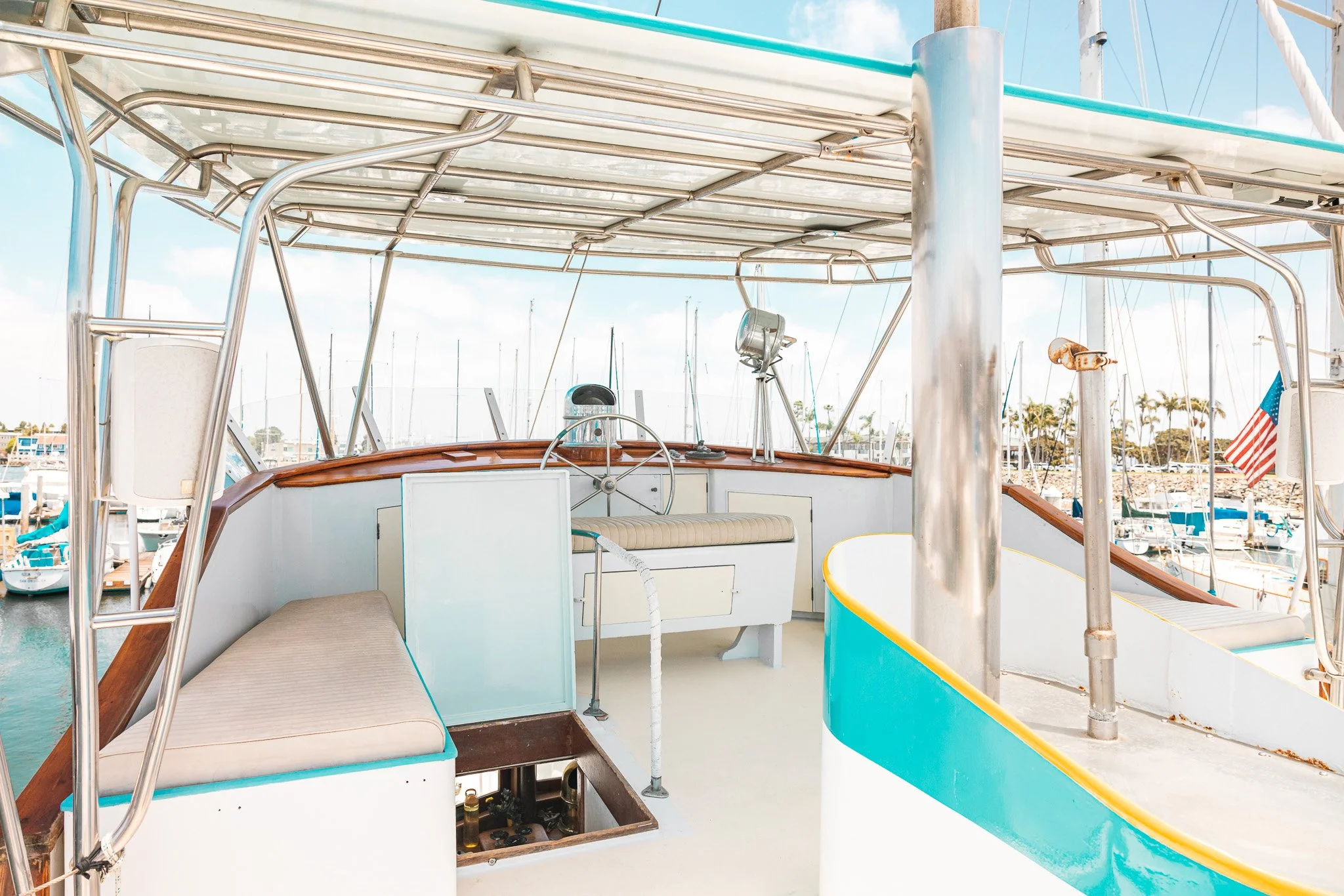 View of the deck of a boat with a beige cushioned seat, steering wheel, and navigation equipment, docked at a marina with sailboats and an American flag in the background under a sunny sky.