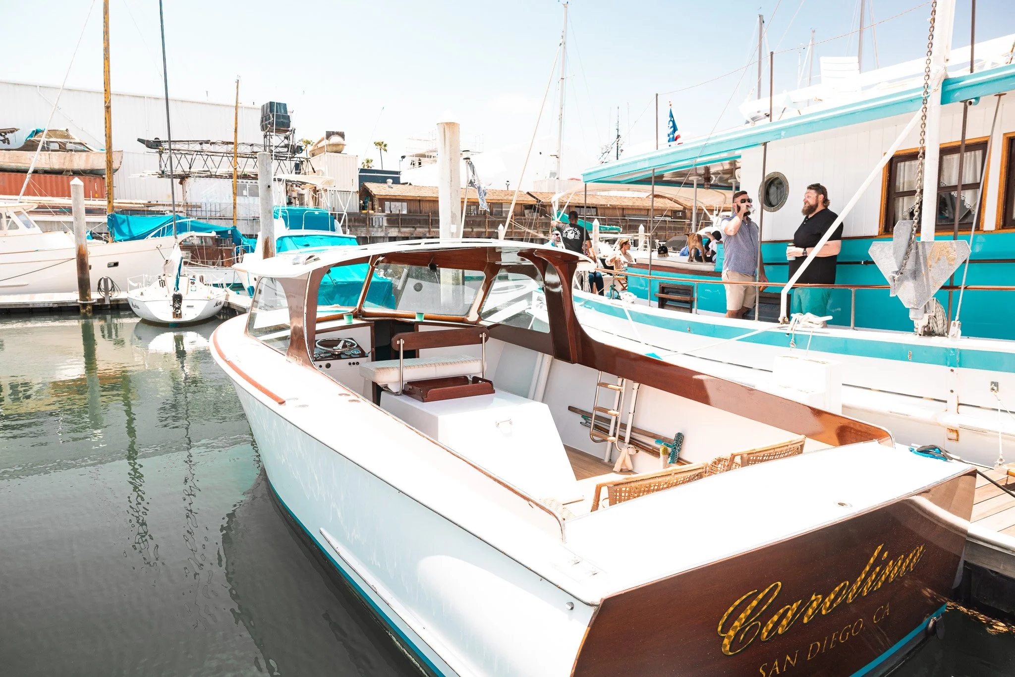 A white and brown yacht named 'Caspian' is docked in a marina, with boats and people in the background.