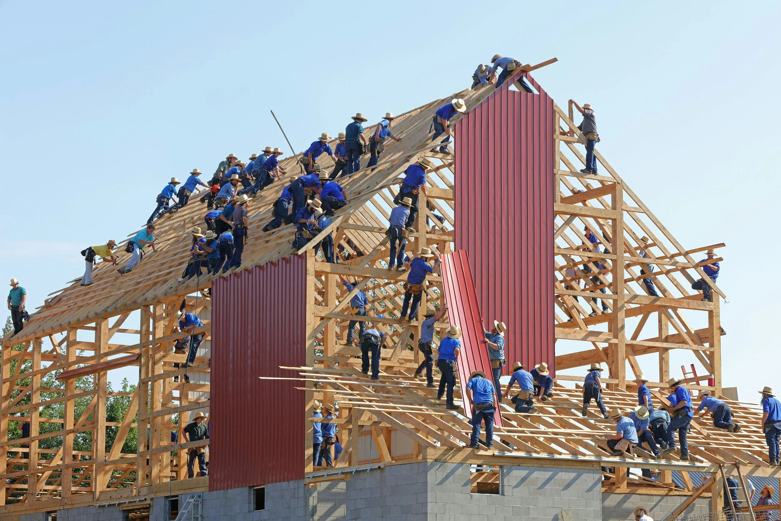 Construction workers building a wooden roof frame, installing red metal panels, on a large building during daytime.