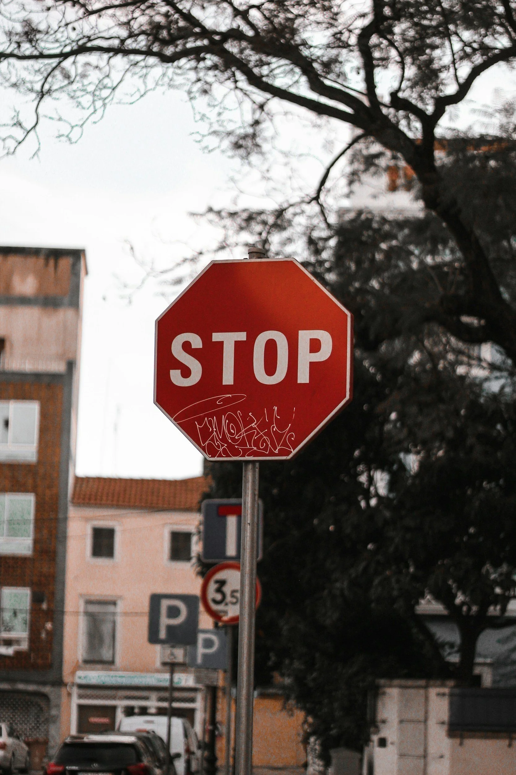 Urban stop sign with graffiti in Setúbal, Portugal, against buildings and tree branches.