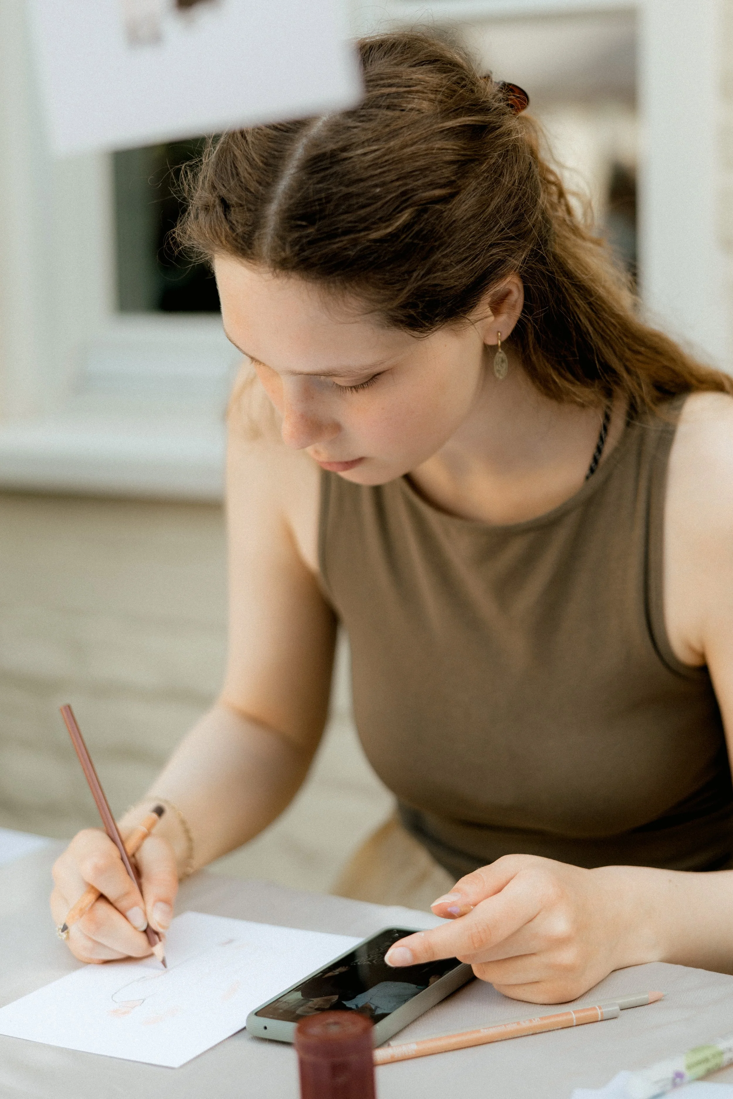 A young woman with brown hair and earrings is sitting at a table, drawing on a piece of paper with a pencil. She is also holding a smartphone in her left hand and has various drawing supplies on the table.