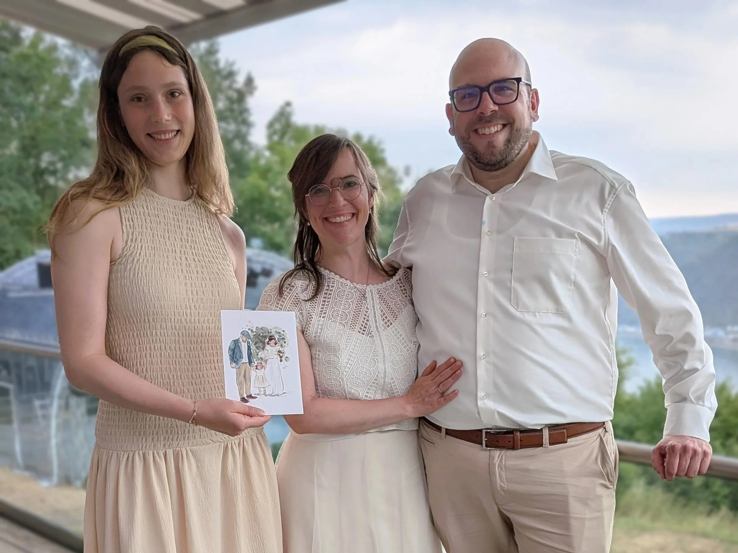 A group of four people standing together outdoors, smiling. One woman is holding a drawing or card of two people walking, with trees and sky in the background.