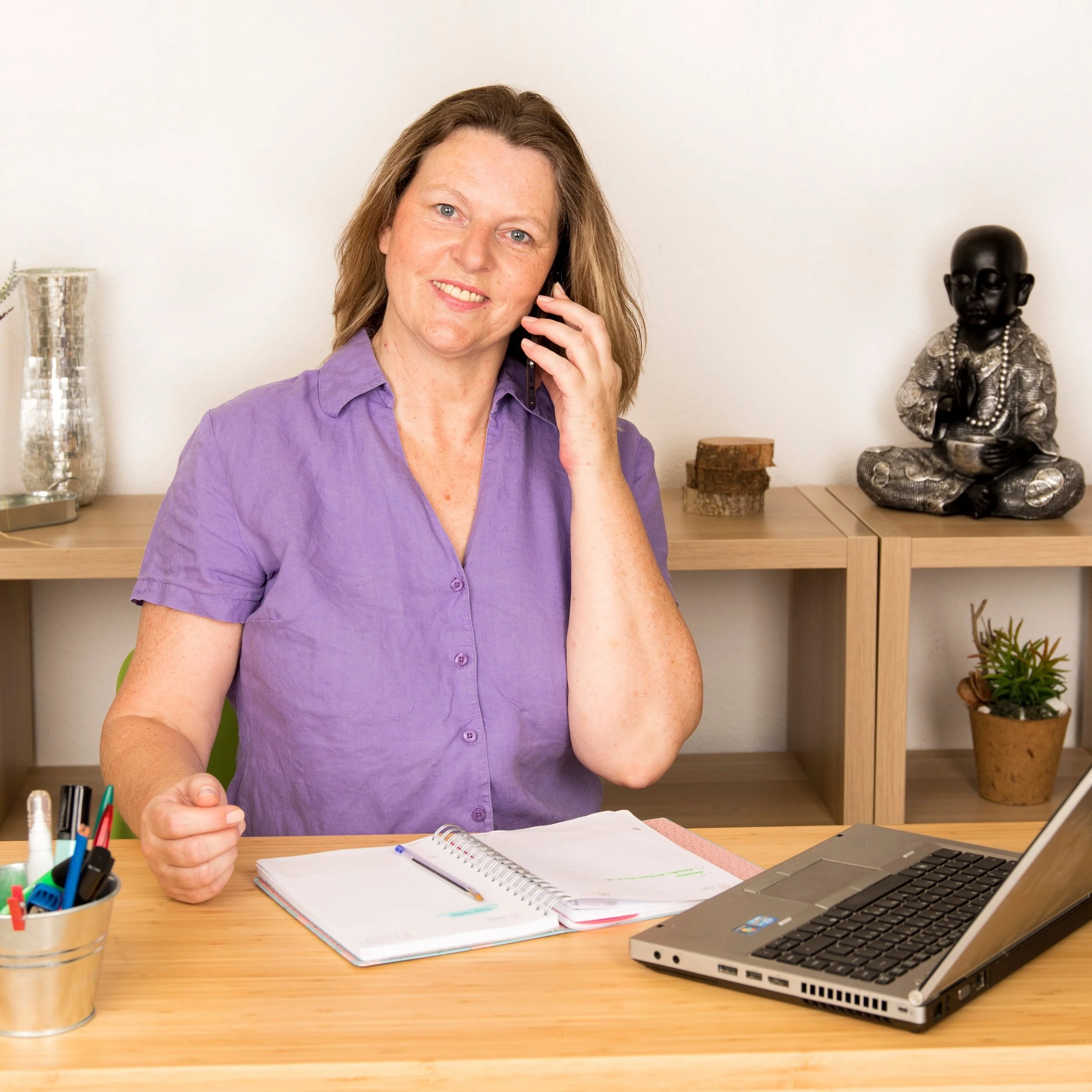 Eine Frau sitzt an einem Schreibtisch und spricht am Telefon. Sie hat einen offenen Notizblock vor sich.