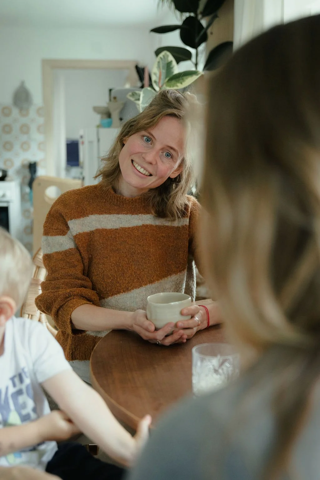 Eine Frau mit lockigem braunem Haar, die ein braun-cremefarbenes gestreiftes Pullover trägt, sitzt an einem Tisch und hält eine weiße Tasse in den Händen. Sie lächelt freundlich und schaut zu einer Person, die nur teilweise im Bild zu sehen ist.