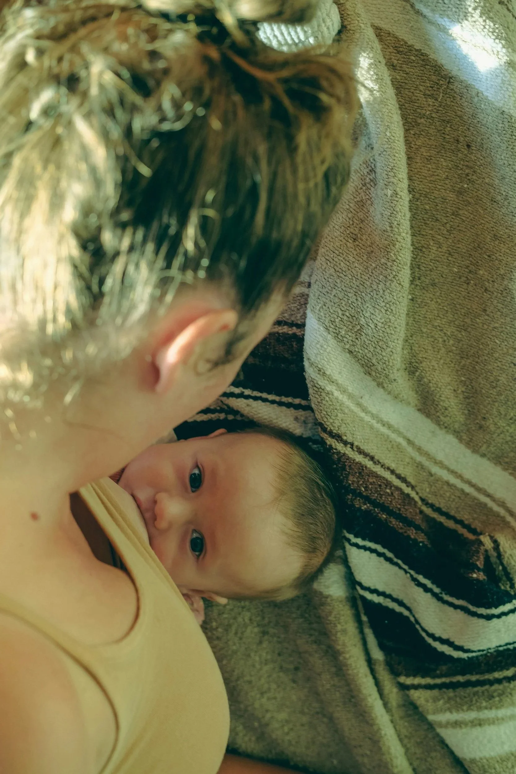 Eine Frau stillt ihr Baby, es liegt auf einer gestreiften Decke und blickt in die Kamera.