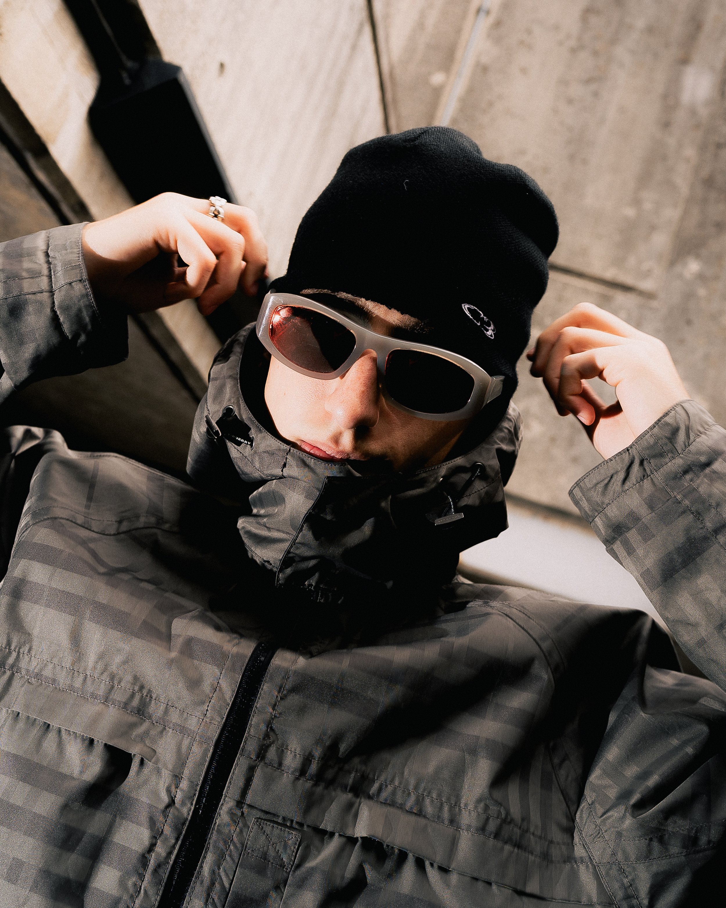 Top-down view of a person wearing black sunglasses, a black beanie, and a gray jacket, adjusting the beanie with both hands. The background shows a gray concrete floor.