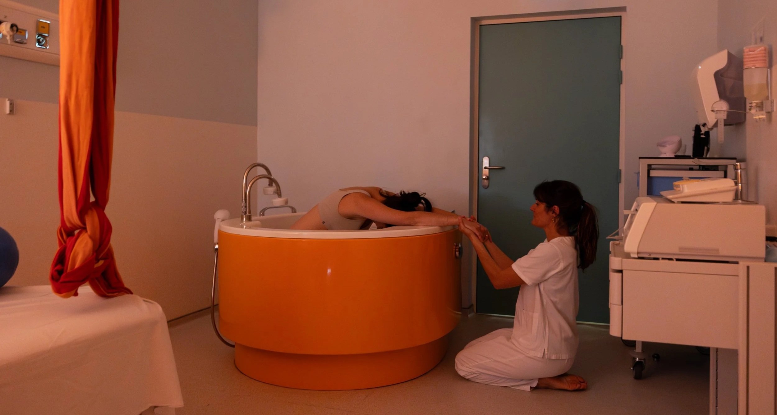 Une femme allongée dans un bain de baignoire orange, une autre femme l'aide en lui tenant la main, dans une salle d'hôpital.