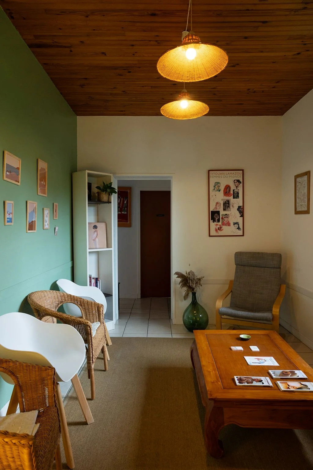 Salon de style cosy avec des chaises en rotin et en plastique blanc, un fauteuil en tissu gris, un grand vase vert avec des fleurs séchées, des tableaux accrochés au mur, et un plafond en bois avec deux lampes en rotin.