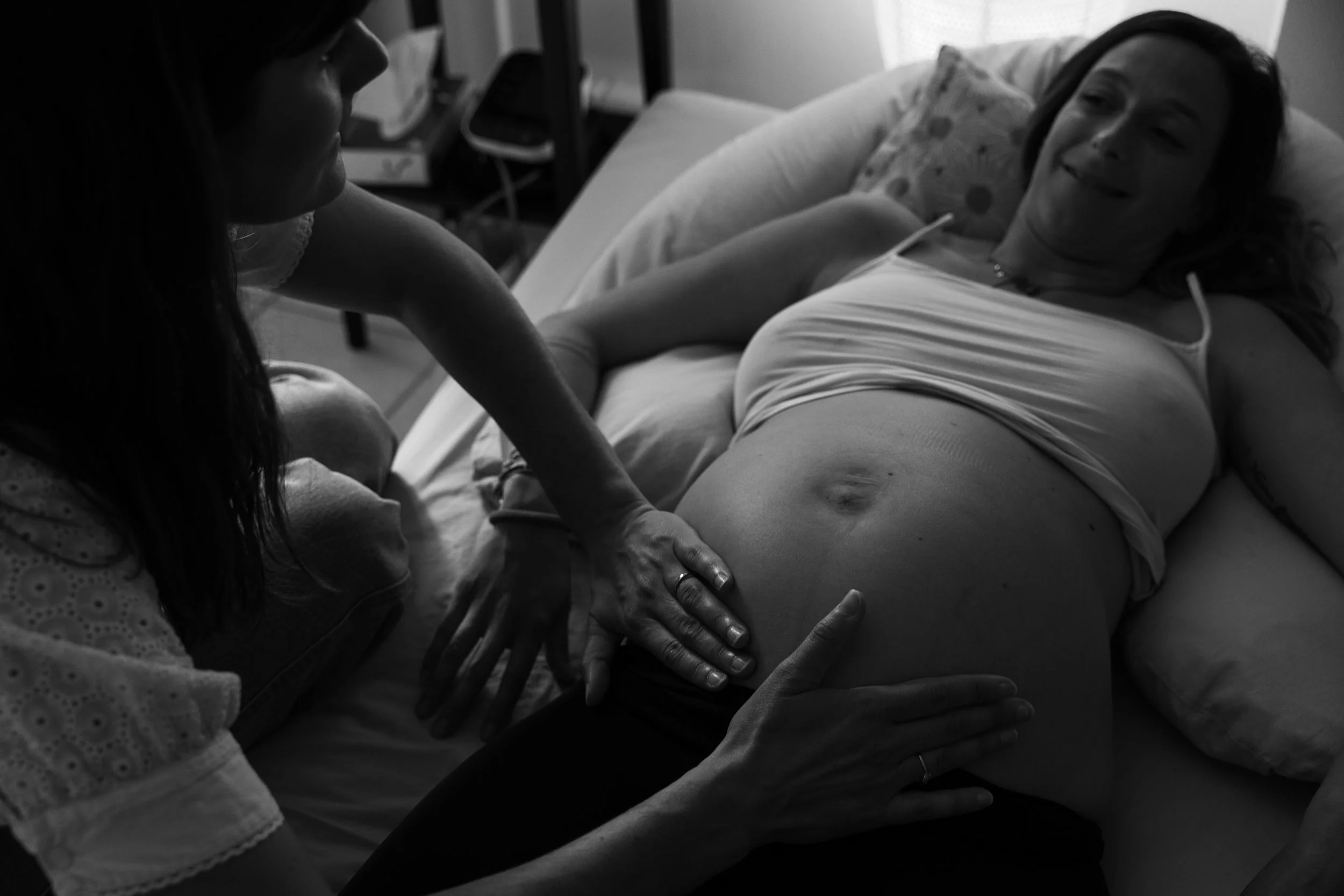 Une femme enceinte est allongée sur un lit dans une pièce, pendant qu'une autre femme, probablement une professionnelle de santé, touche son ventre et son ventre d'une main. La femme allongée sourit, une autre main repose sur son ventre.