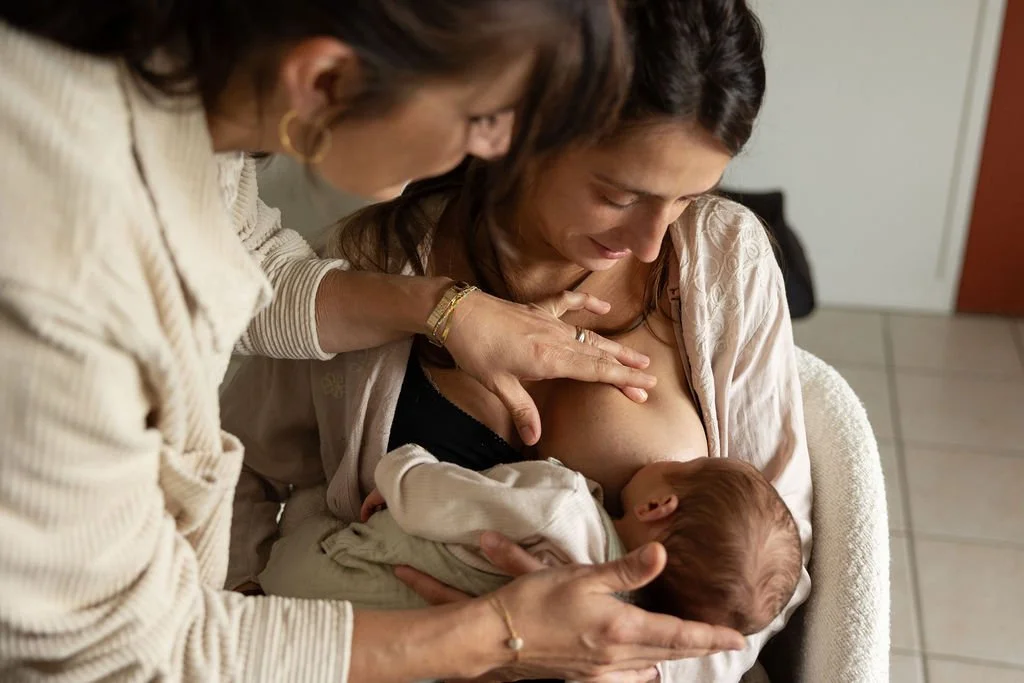 Une femme enceinte allaite son bébé, avec une sage-femme qui l'aide, dans un environnement intérieur.