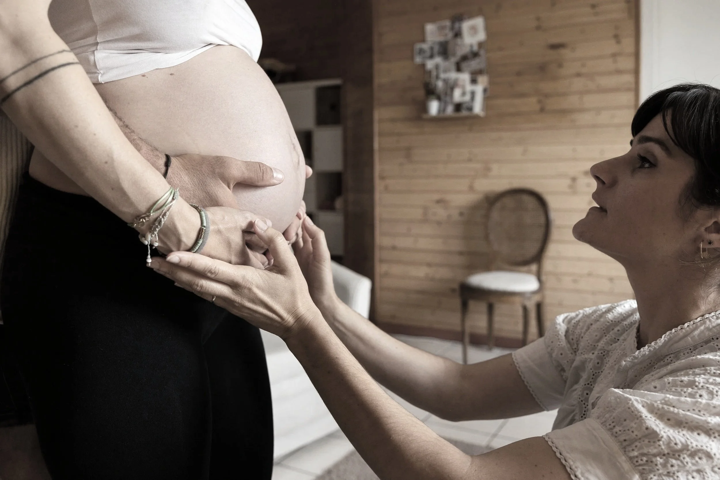 Une femme enceinte tenant le ventre d'une autre femme, qui semble la soutenir ou parler avec elle dans une pièce avec un mur en bois et une chaise en arrière-plan.